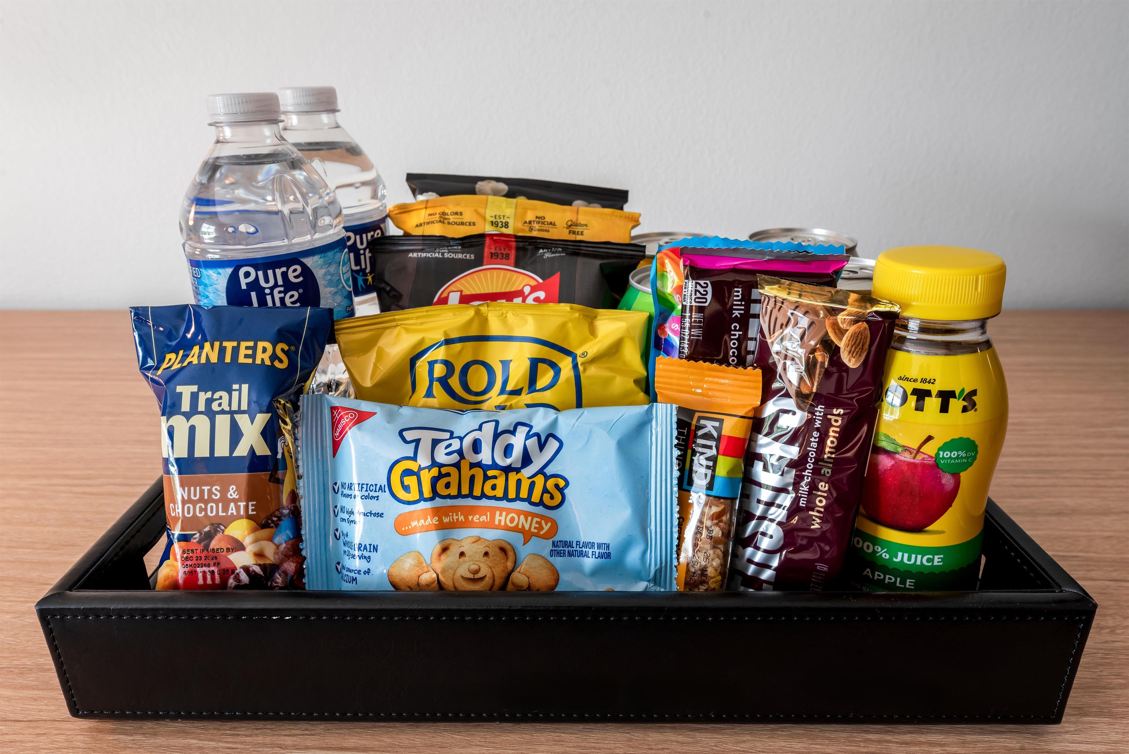 A black tray containing various food items including Pure Life water bottles, Trail Mix, Rold Teddy Grahams, milk chocolate with almonds, and a bottle of apple juice.