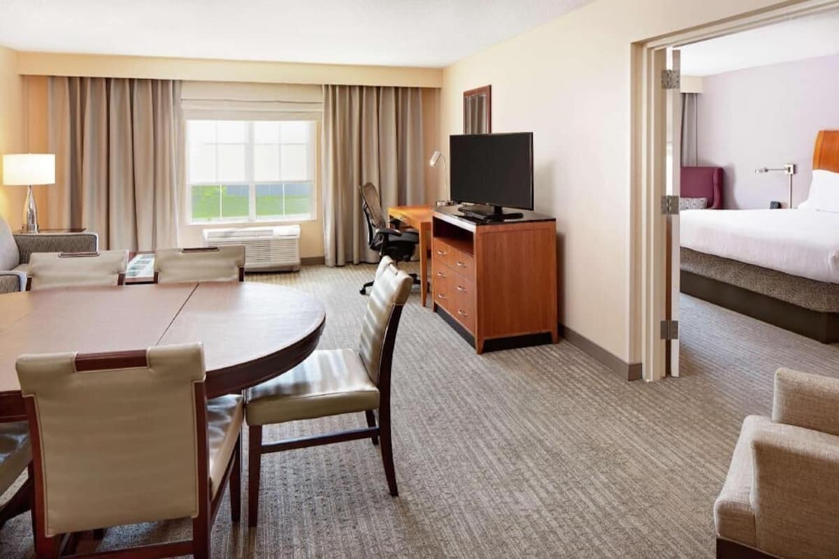 A hotel room with a dining table, chairs, a TV, a bed, and a view of the outdoors.