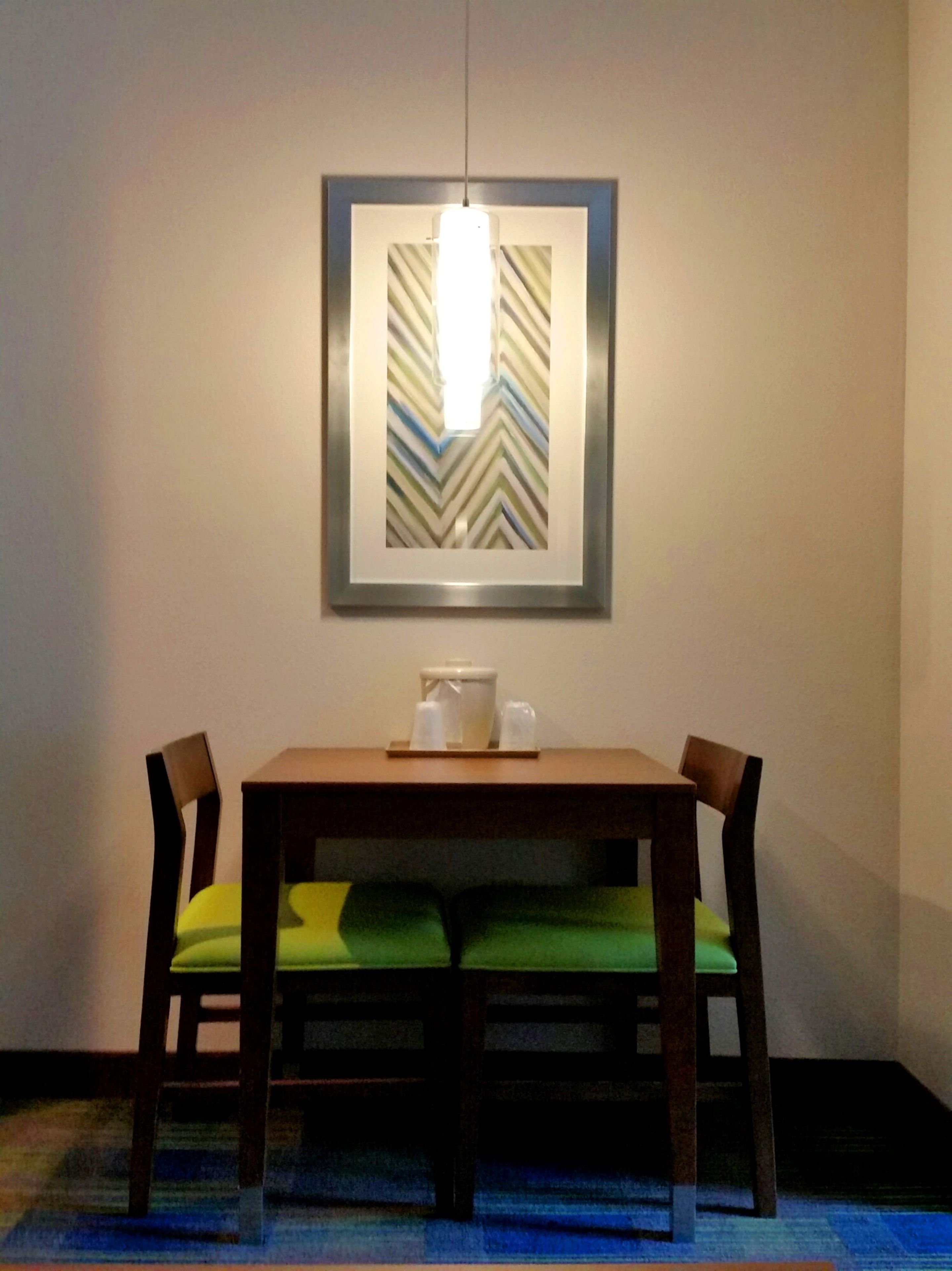 A dining area with a table and chairs, a framed artwork above the table, and a hanging light fixture.