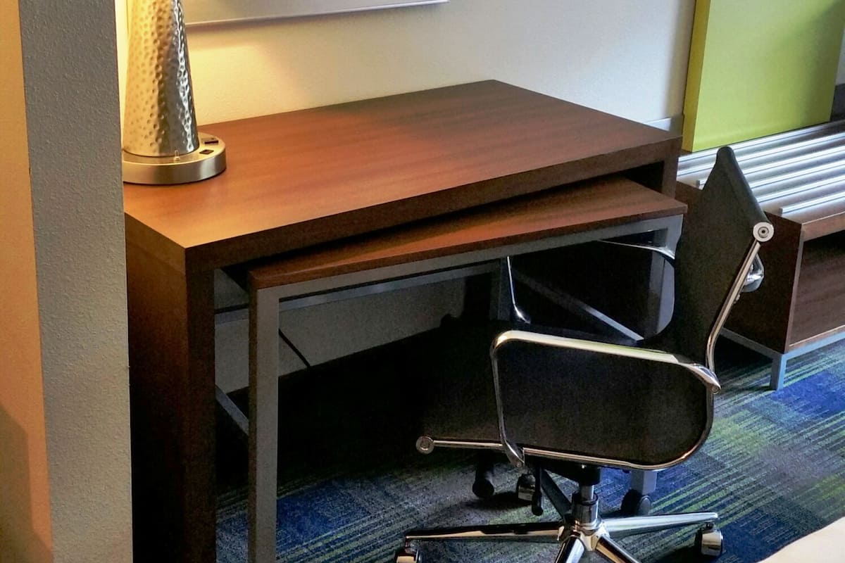 A hotel room with a wooden desk, a chair, a lamp, and a bed.
