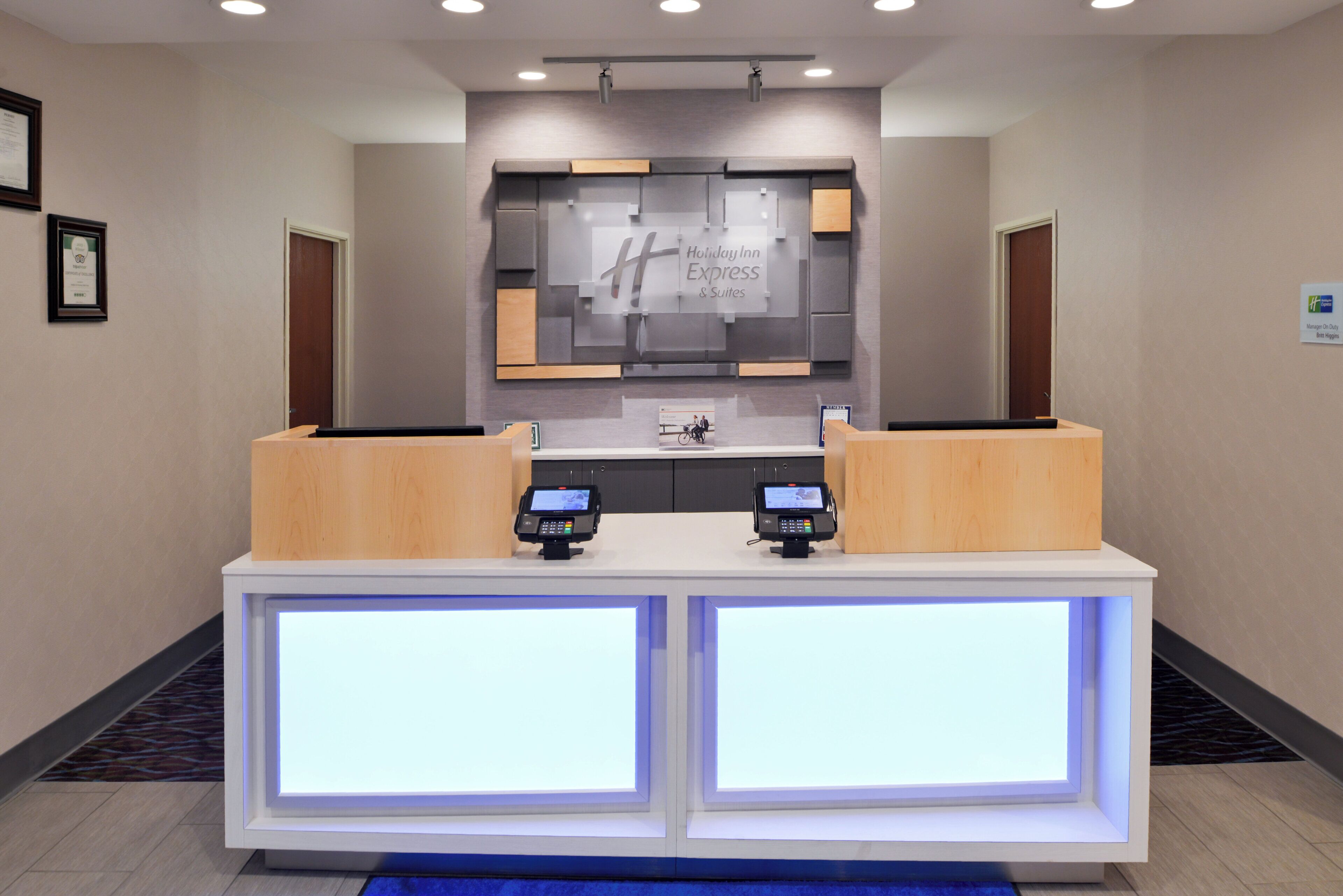 A reception desk with two digital kiosks, a large illuminated sign for Holiday Inn Express & Suites, and framed certificates on the wall.