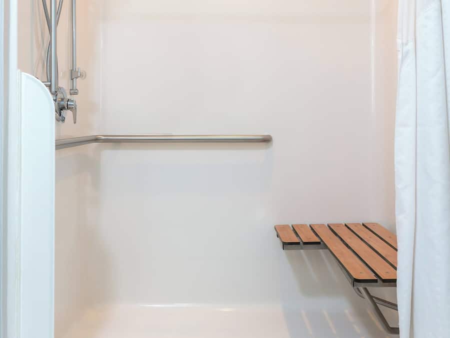 Cabina de ducha con puerta de cristal, cabezal de ducha metálico, ducha de mano, barra de apoyo metálica y banco de madera.