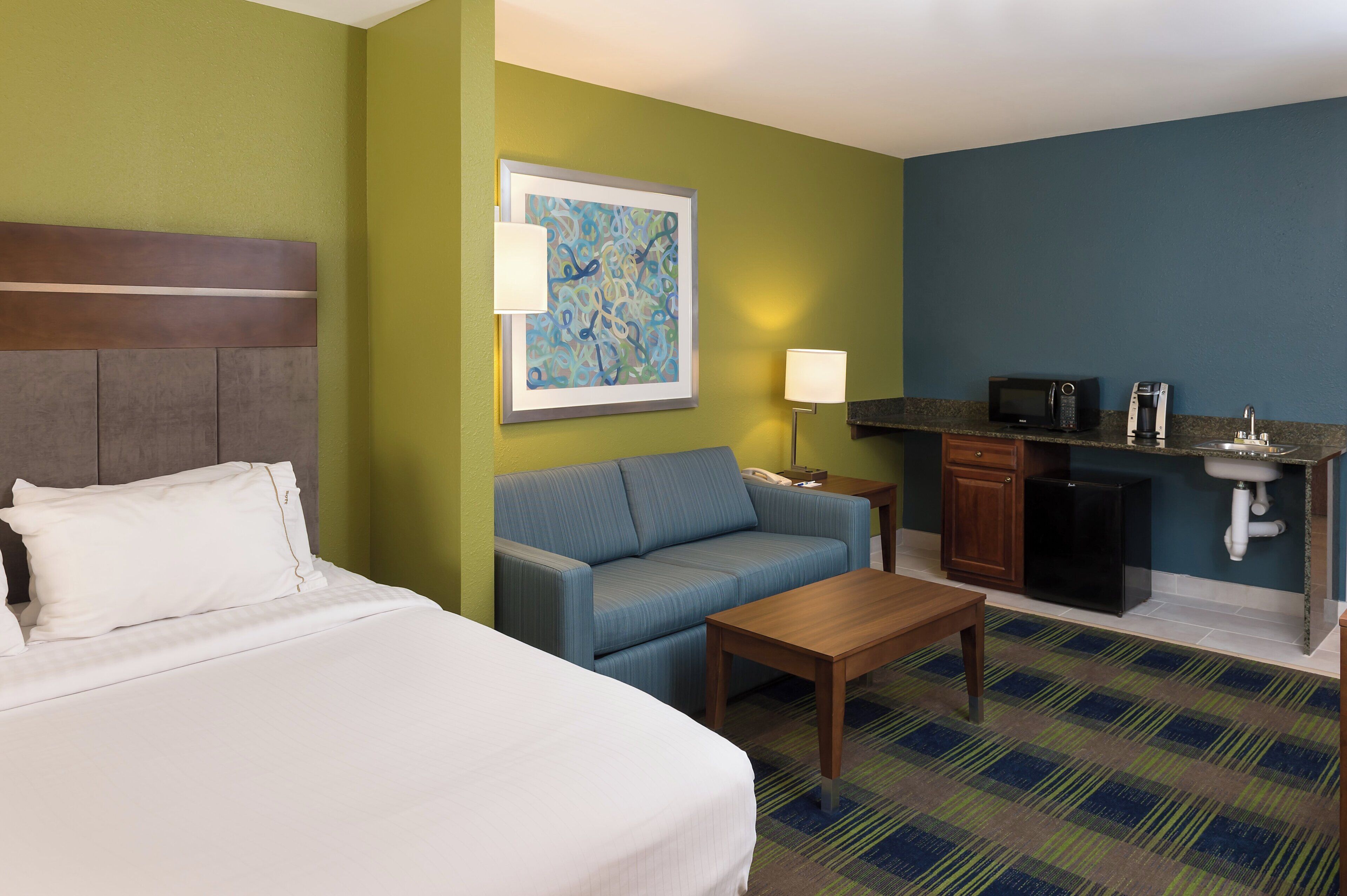 A hotel room with a bed, a sofa, a coffee table, a desk, a microwave, a refrigerator, and a sink.