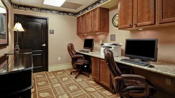 A hotel room with a desk, two chairs, a computer, a television, and a clock on the wall.