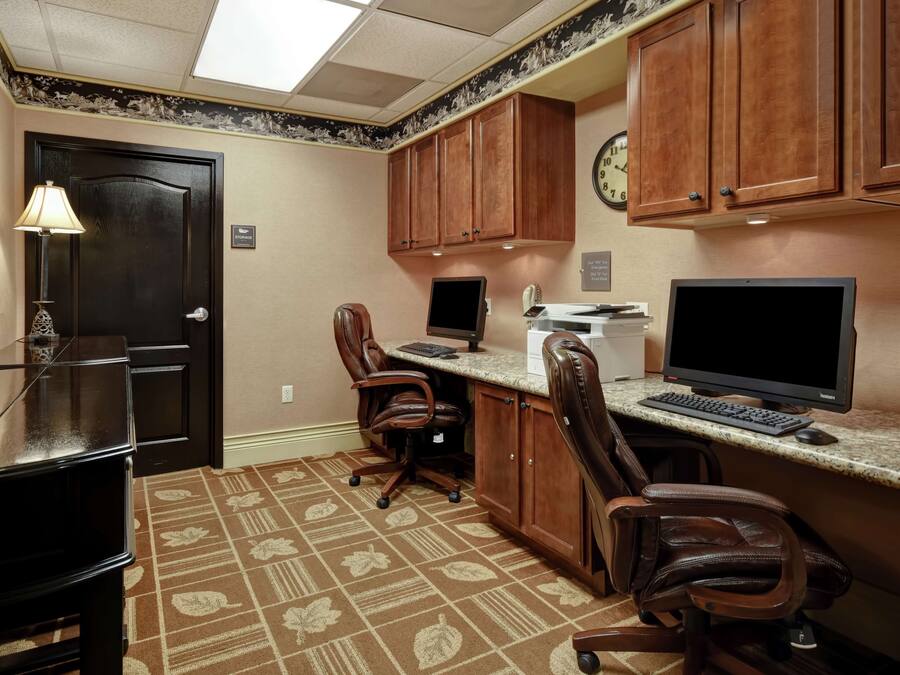 A hotel room with a desk, two chairs, a computer, a television, and a clock on the wall.