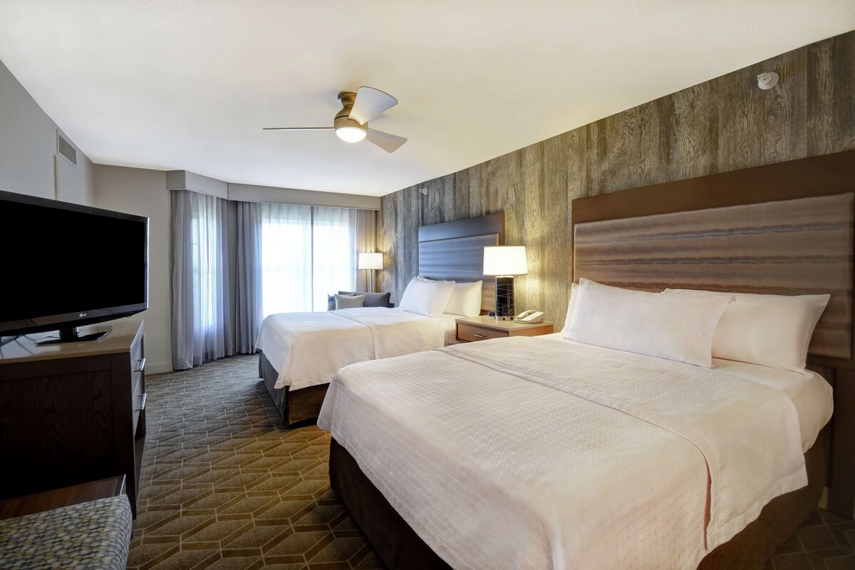 Habitación de hotel con dos camas, televisión, ventilador de techo y un gran ventanal con cortinas.