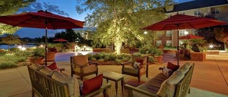Un área de estar al aire libre con muebles de madera y sombrillas rojas, rodeada de plantas bien iluminadas y un edificio al fondo.