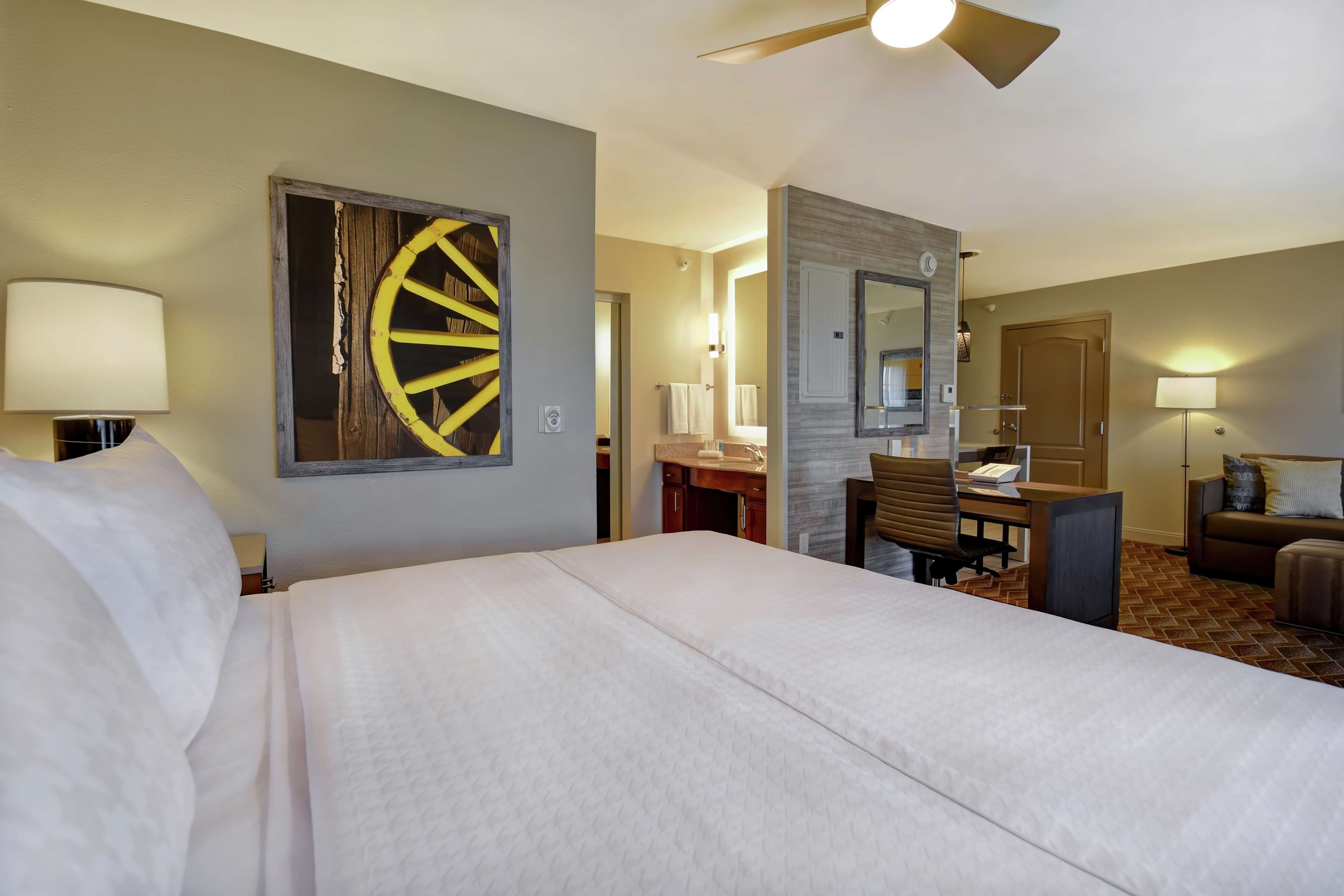 Ein Hotelzimmer mit einem großen Bett, einem Schreibtisch mit Stuhl, einer Schreibtischlampe, einem Wandspiegel und einem Deckenventilator.