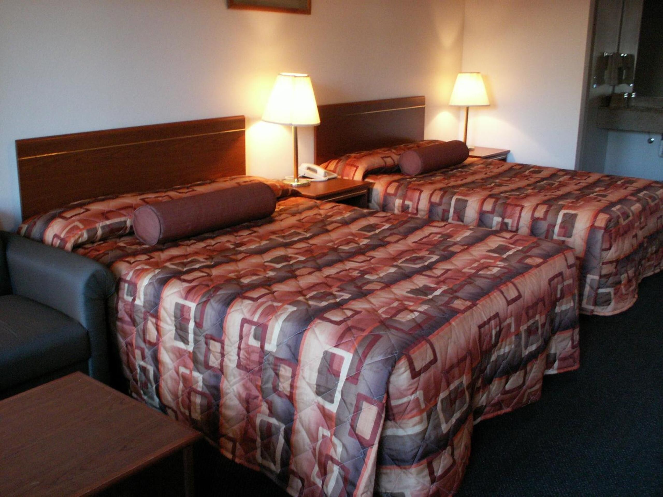 A hotel room with two beds, each with a patterned bedspread, a nightstand, a lamp, and a chair.