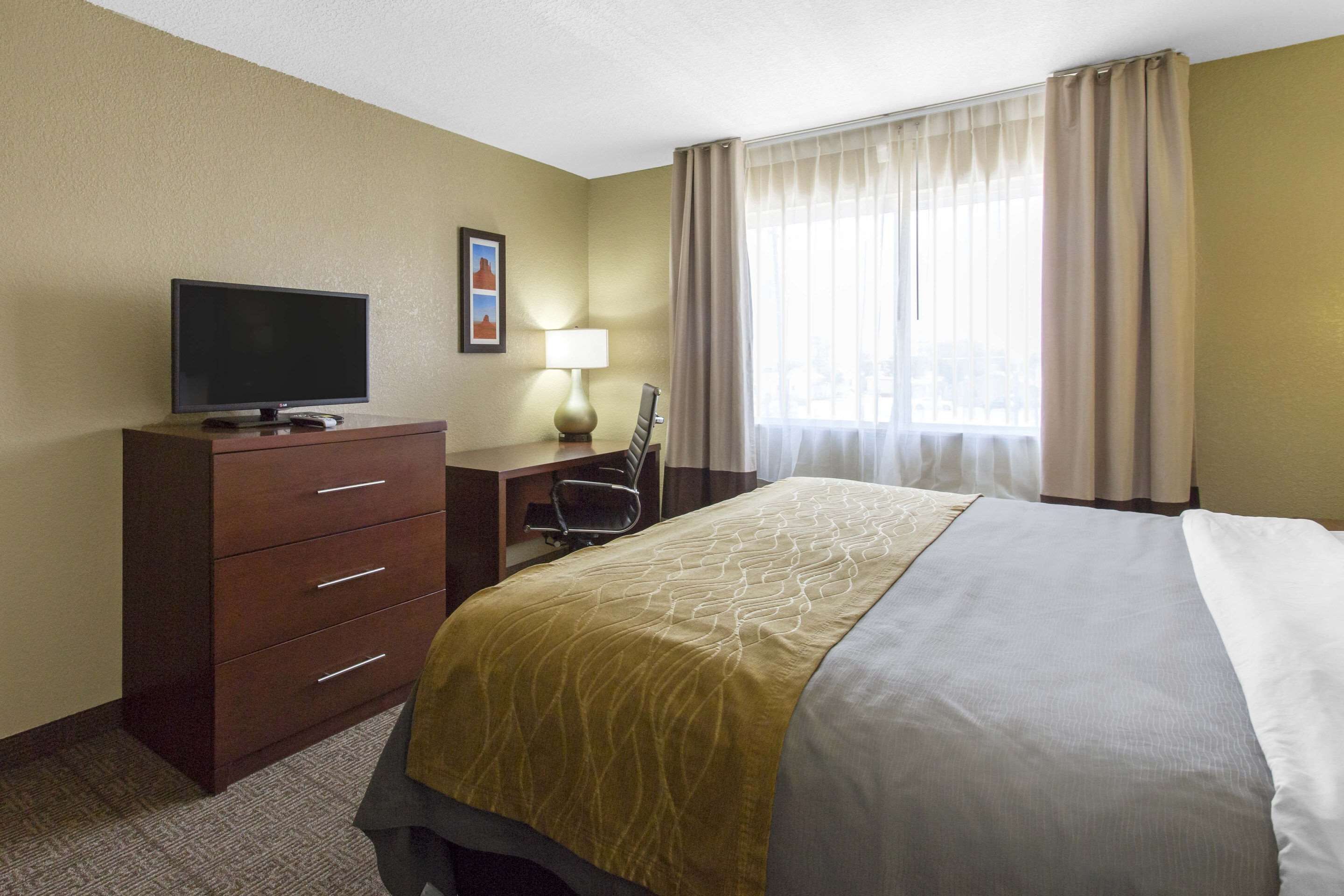 A hotel room with a bed, a desk with a computer, a TV, a lamp, and a window with curtains.