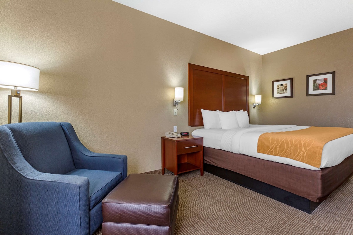 A hotel room with a bed, a blue armchair, a wooden nightstand, and a brown ottoman.