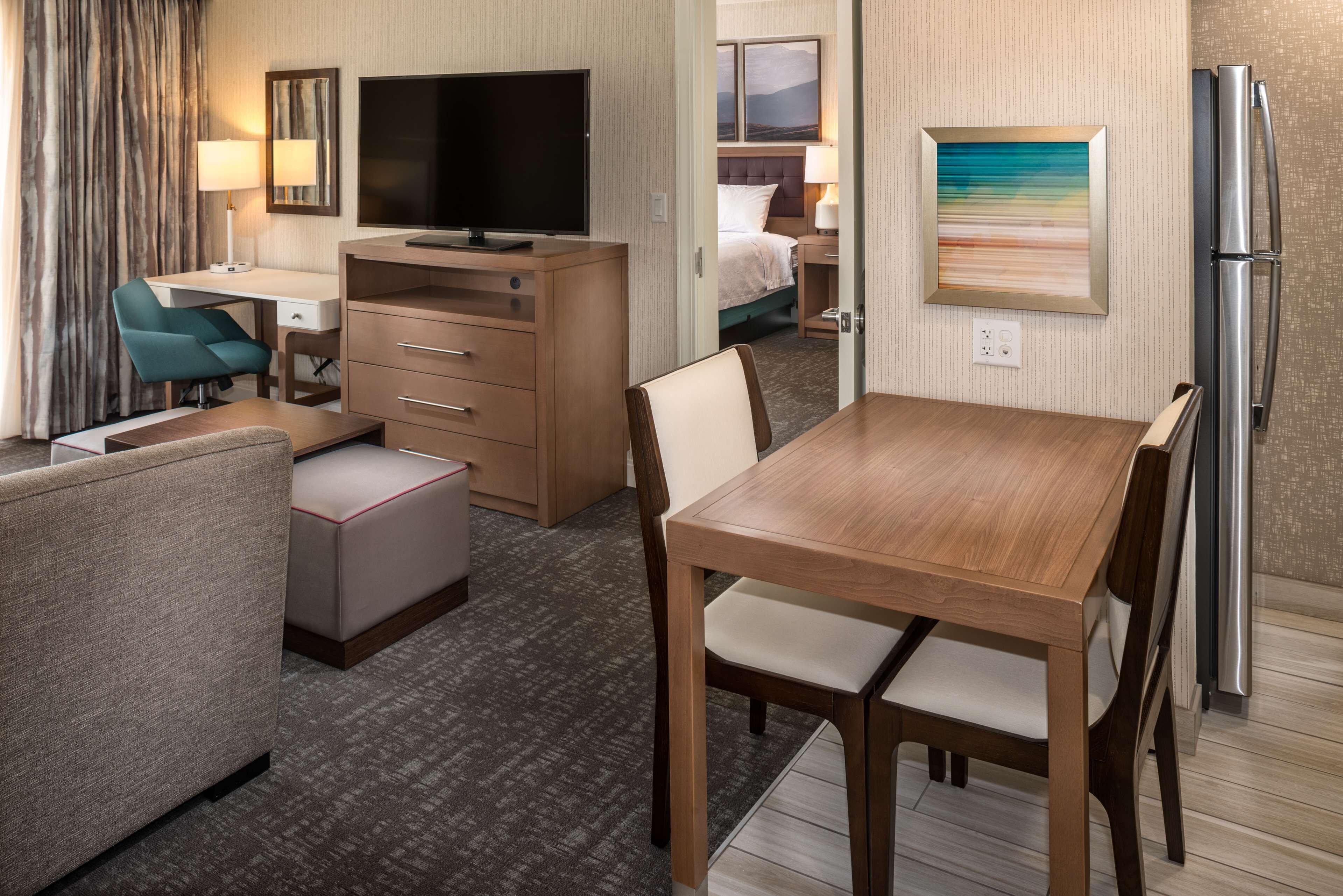 A hotel room with a wooden dining table, a TV, a desk with a chair, a bed, and a refrigerator.