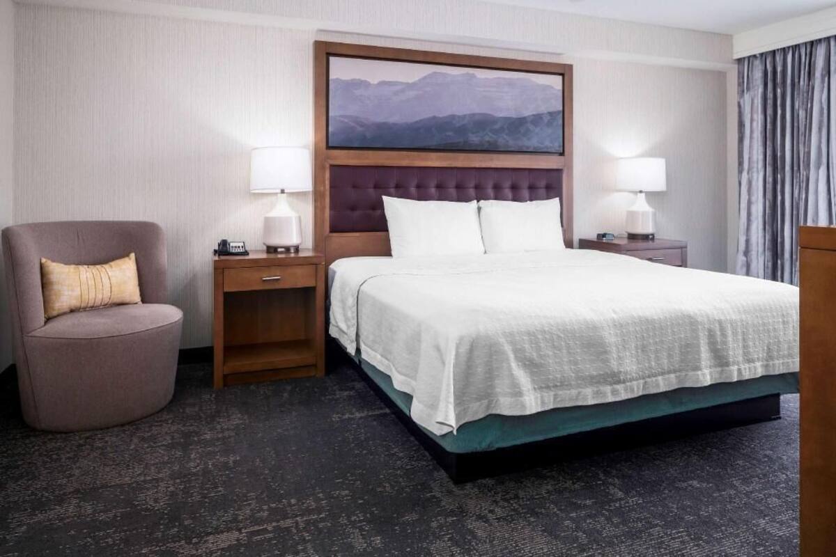Una camera d'albergo con un letto, una sedia, un comodino, una lampada e un quadro appeso al muro.