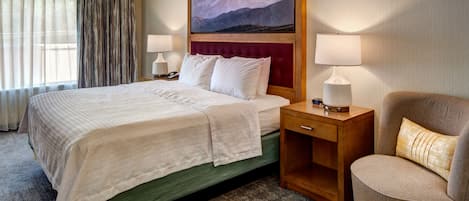 Habitación de hotel con una cama grande, dos mesitas de noche, una silla, un ventilador de techo y un cuadro de un paisaje montañoso en la pared.