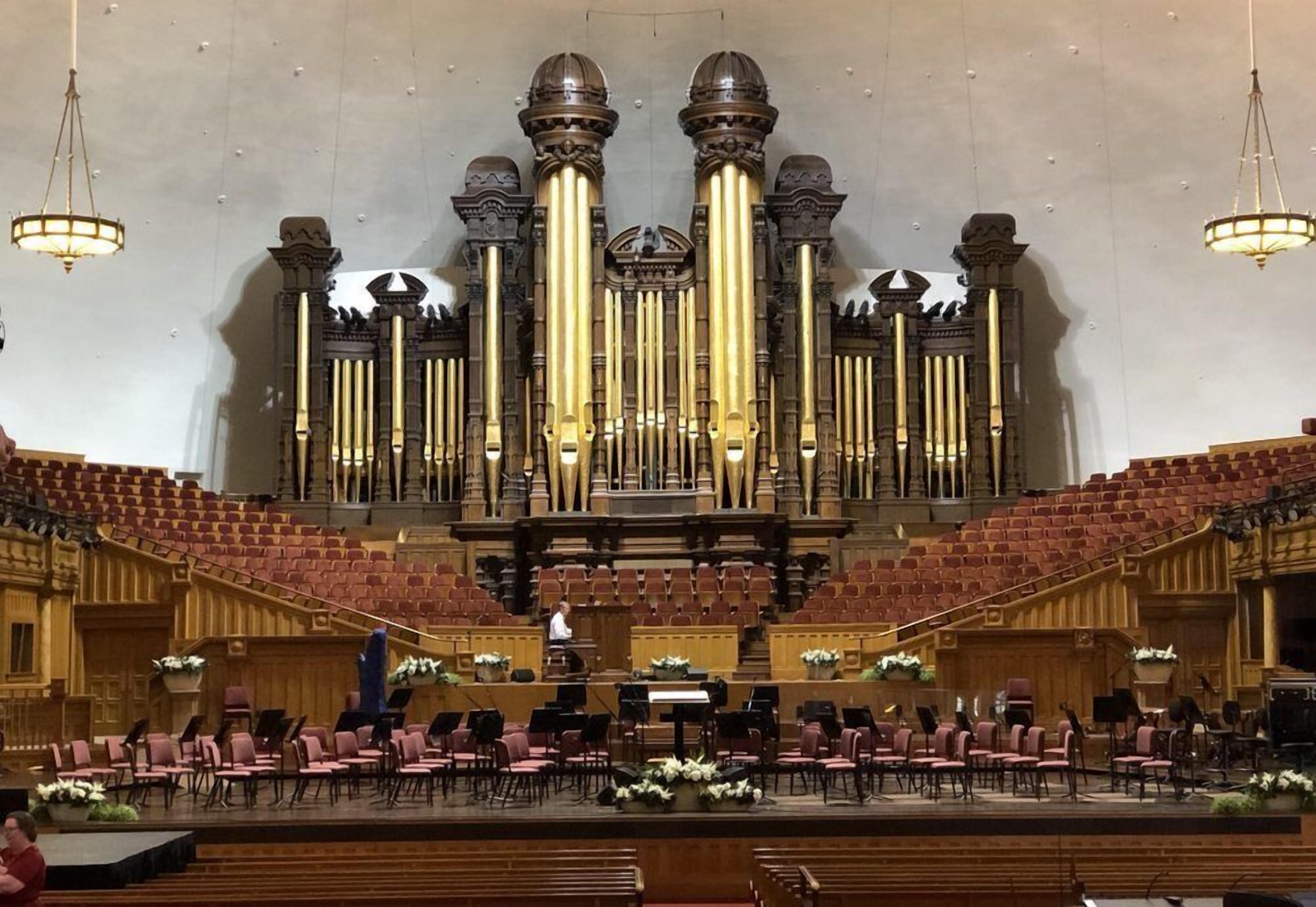 Um órgão com várias tubas e um palco com cadeiras e um púlpito.