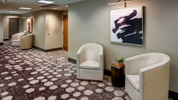 A hotel lobby with a patterned carpet, two armchairs, a framed artwork, and a small plant on a side table.