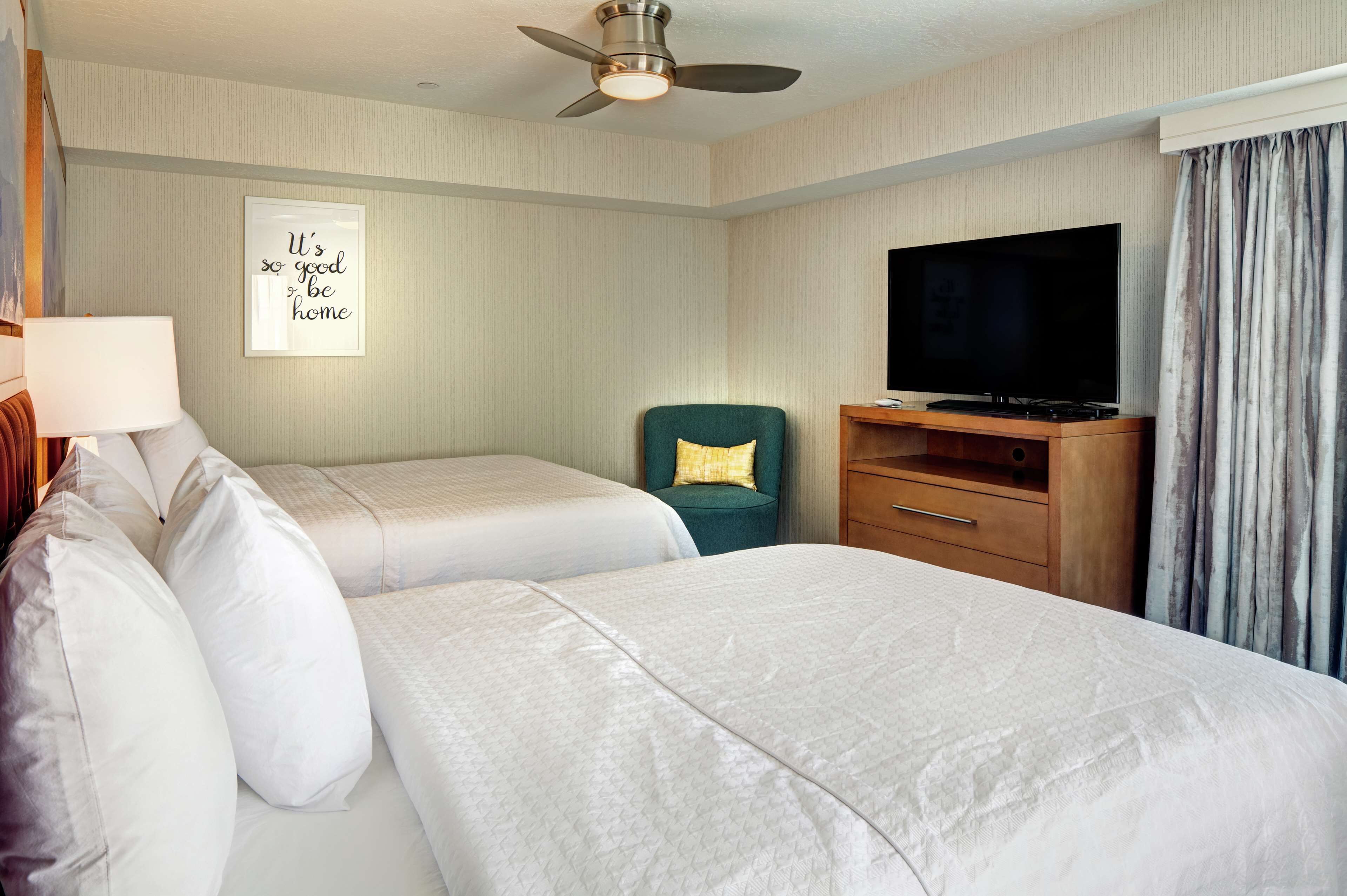 Habitación de hotel con dos camas, televisión, ventilador de techo y una cita enmarcada en la pared.