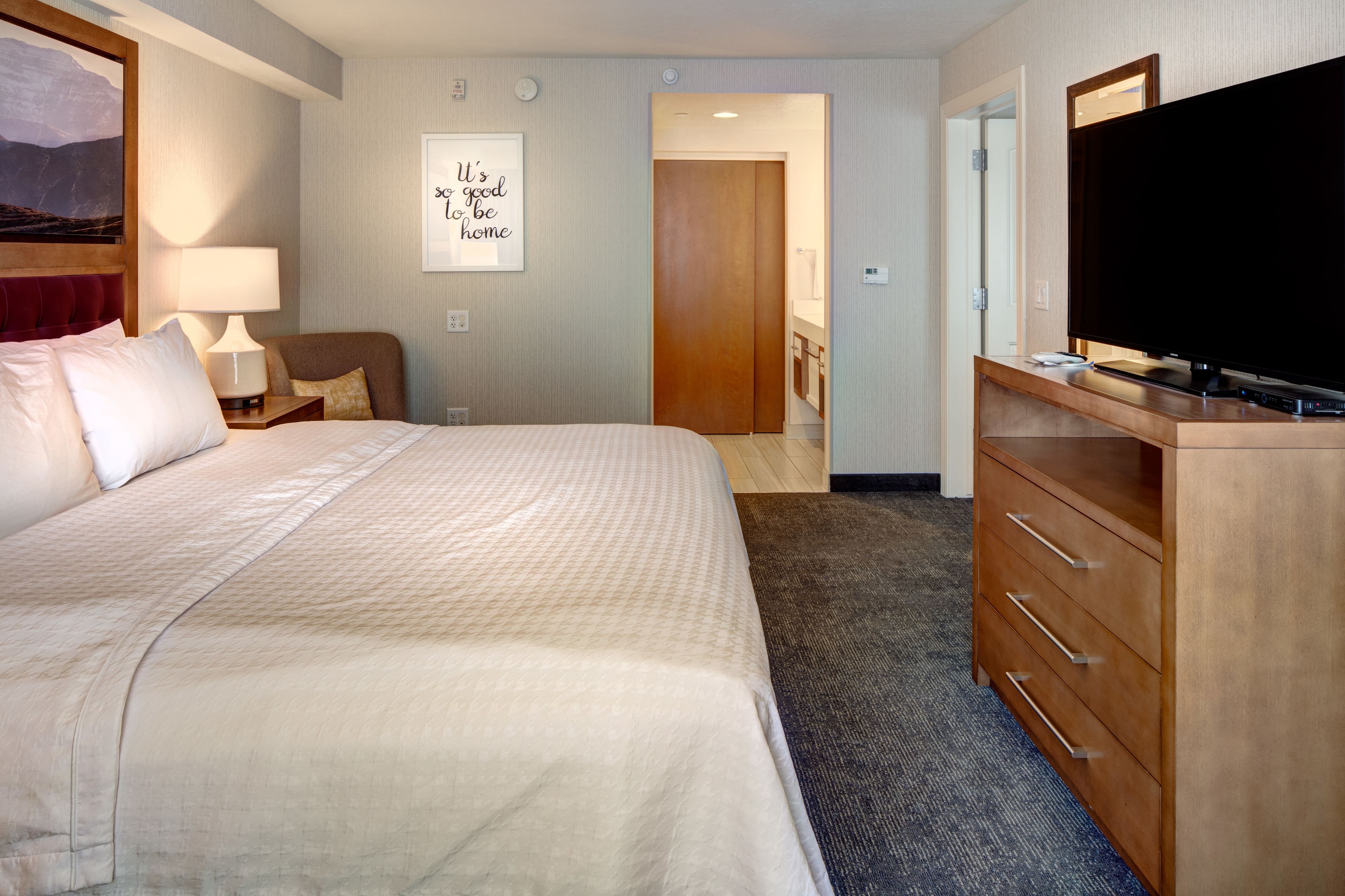 A hotel room with a large bed, a TV, a wooden dresser, a desk, and a bathroom visible through an open door.