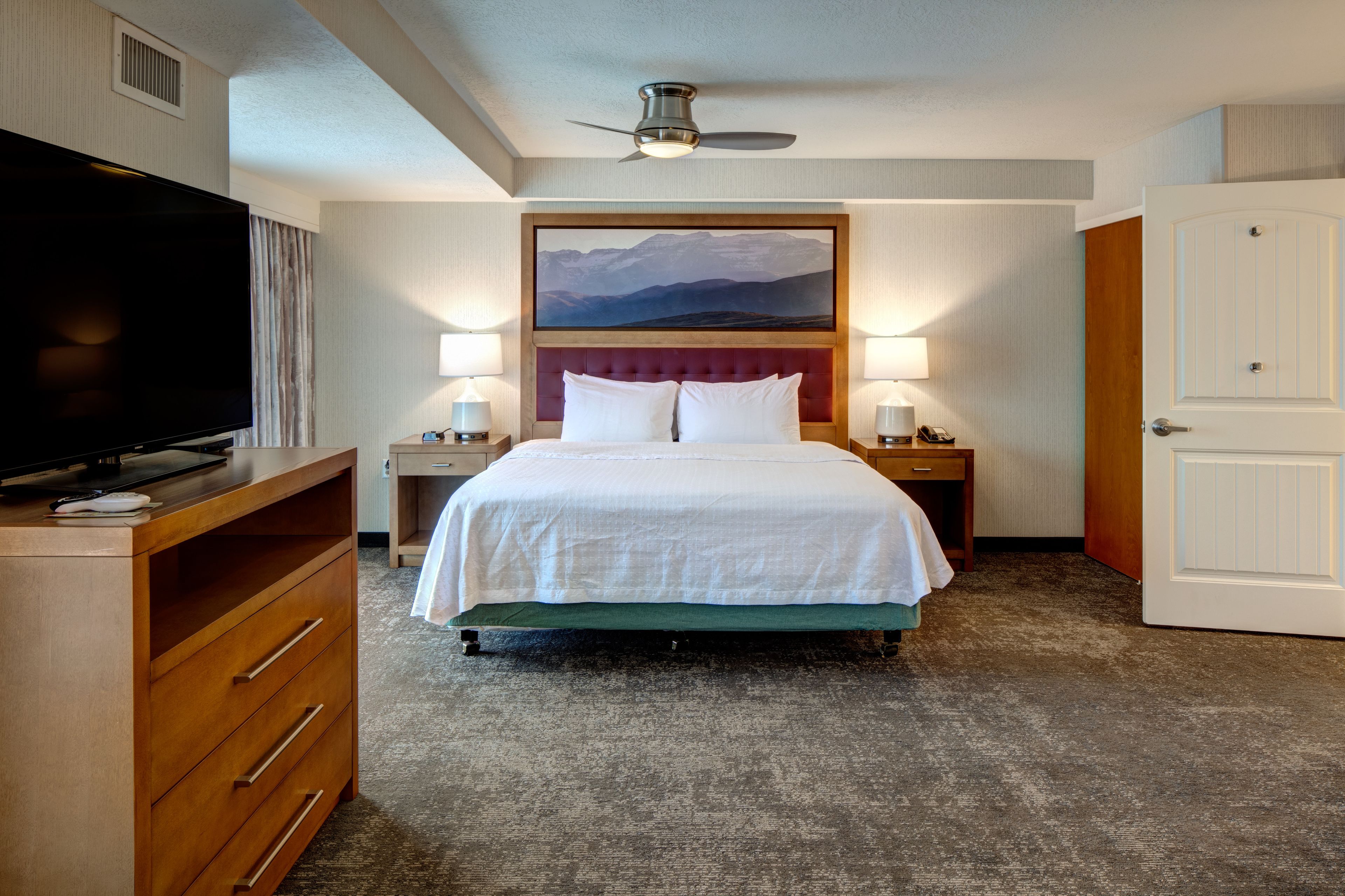 Quarto de hotel com cama, televisão, duas luminárias de cabeceira, um quadro acima da cama e uma porta à direita.