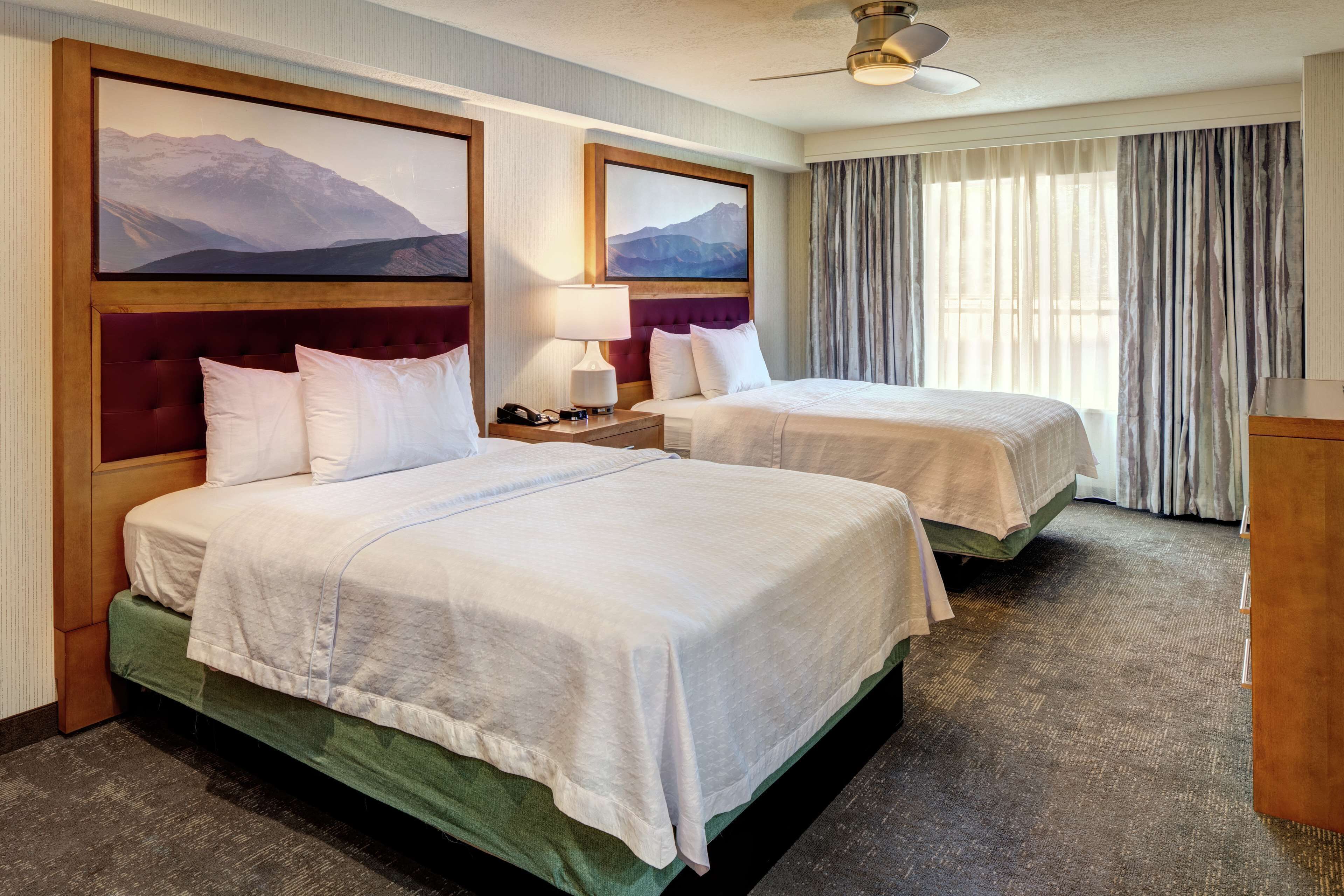A hotel room with two beds, a wooden headboard, a nightstand, a lamp, and a view of mountains through the window.