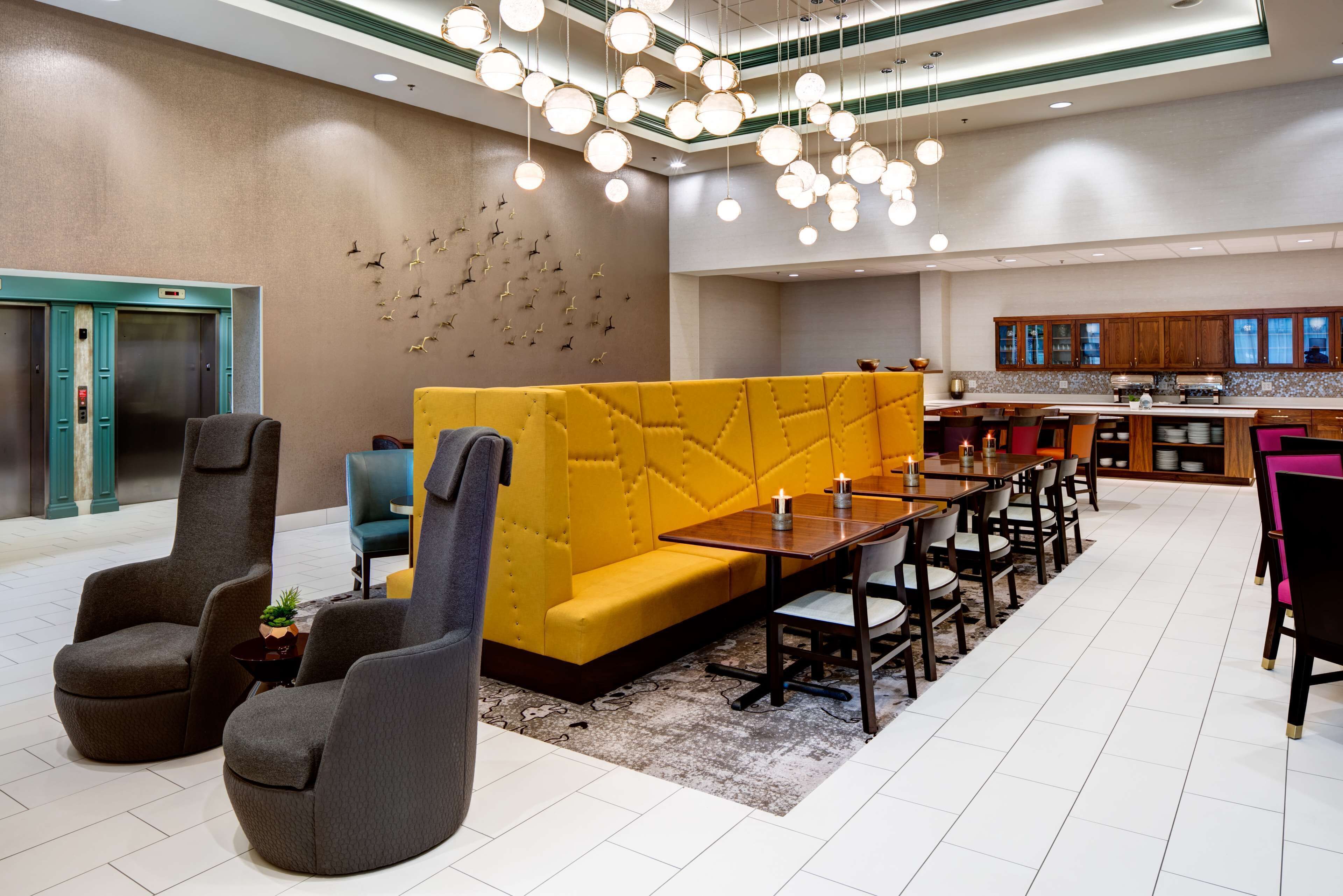 Un moderno vestíbulo de hotel con un sofá amarillo, sillones grises, una mesa de comedor de madera y una cocina al fondo.