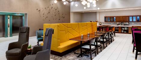 Un moderno vestíbulo de hotel con un sofá amarillo, sillones grises, una mesa de comedor de madera y una cocina al fondo.