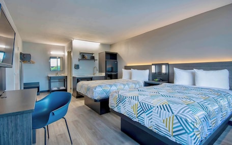 A modern hotel room with a large bed, a desk, a chair, and a small kitchenette.
