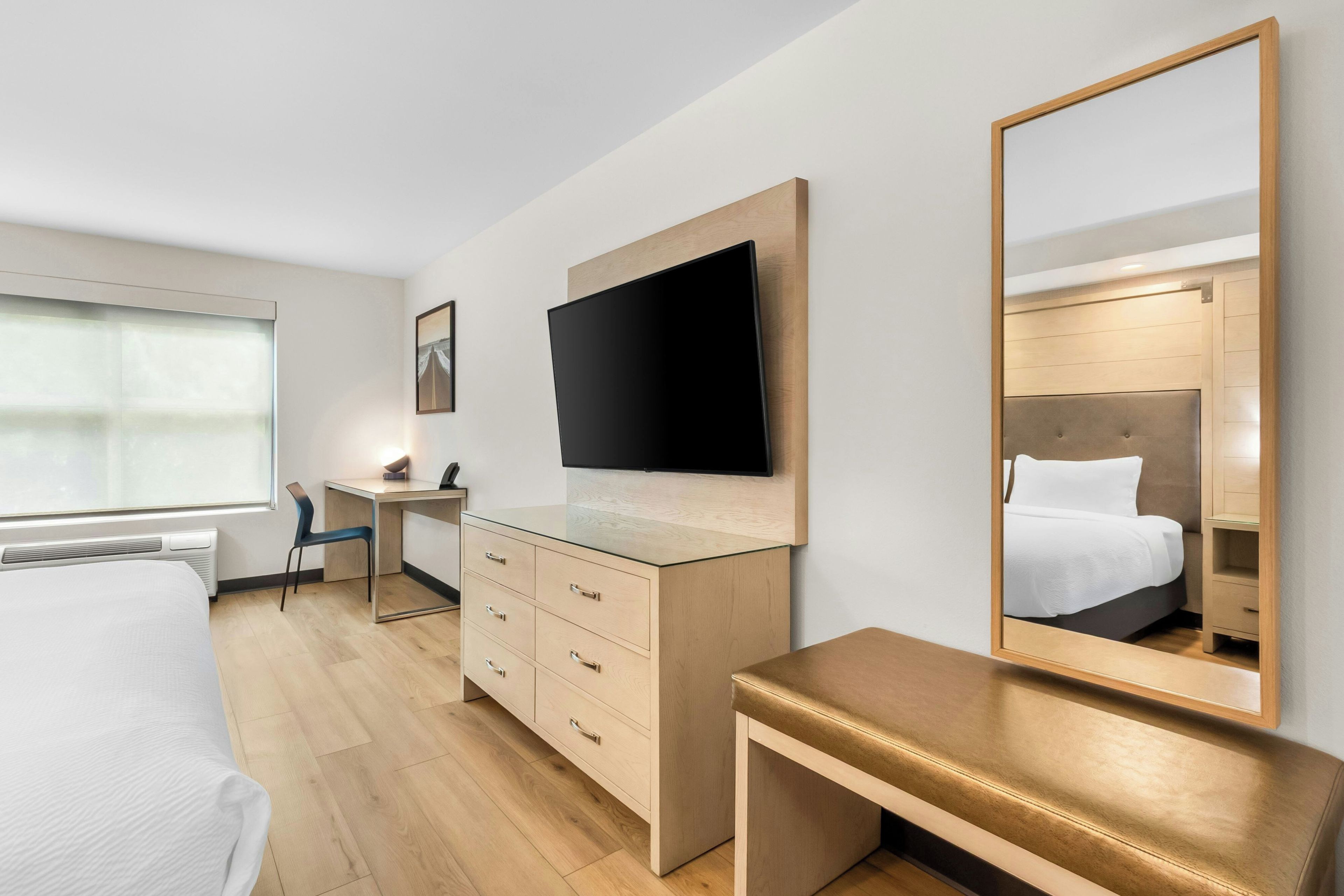 Habitación de hotel con cama, escritorio, silla, televisión, cómoda y un espejo grande.