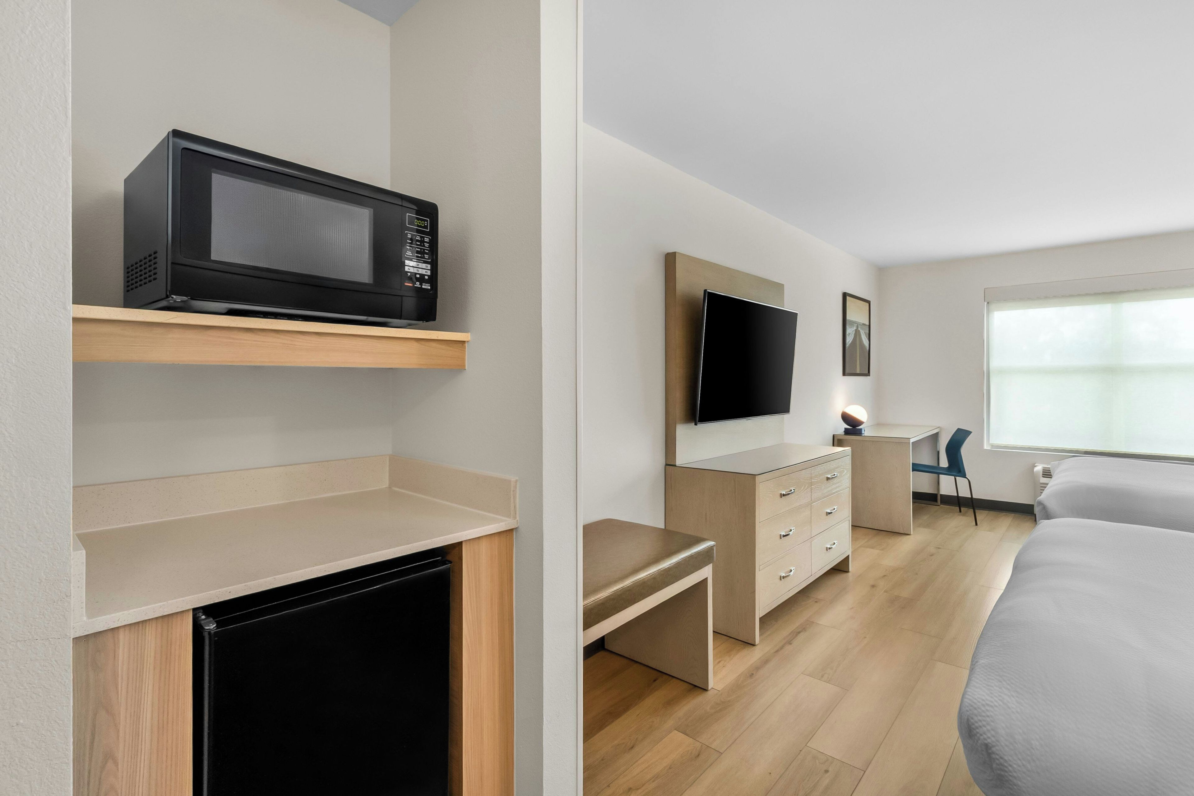 Une chambre d’hôtel moderne équipée d’un téléviseur à écran plat fixé au mur, d’un four à micro-ondes posé sur une étagère, d’un petit bureau avec des tiroirs et d’un lit recouvert d’une couette grise.