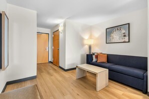 Hotellihuoneessa on puulattia, sohva, sohvapöytä, lamppu ja seinällä oleva kehystetty taulu.