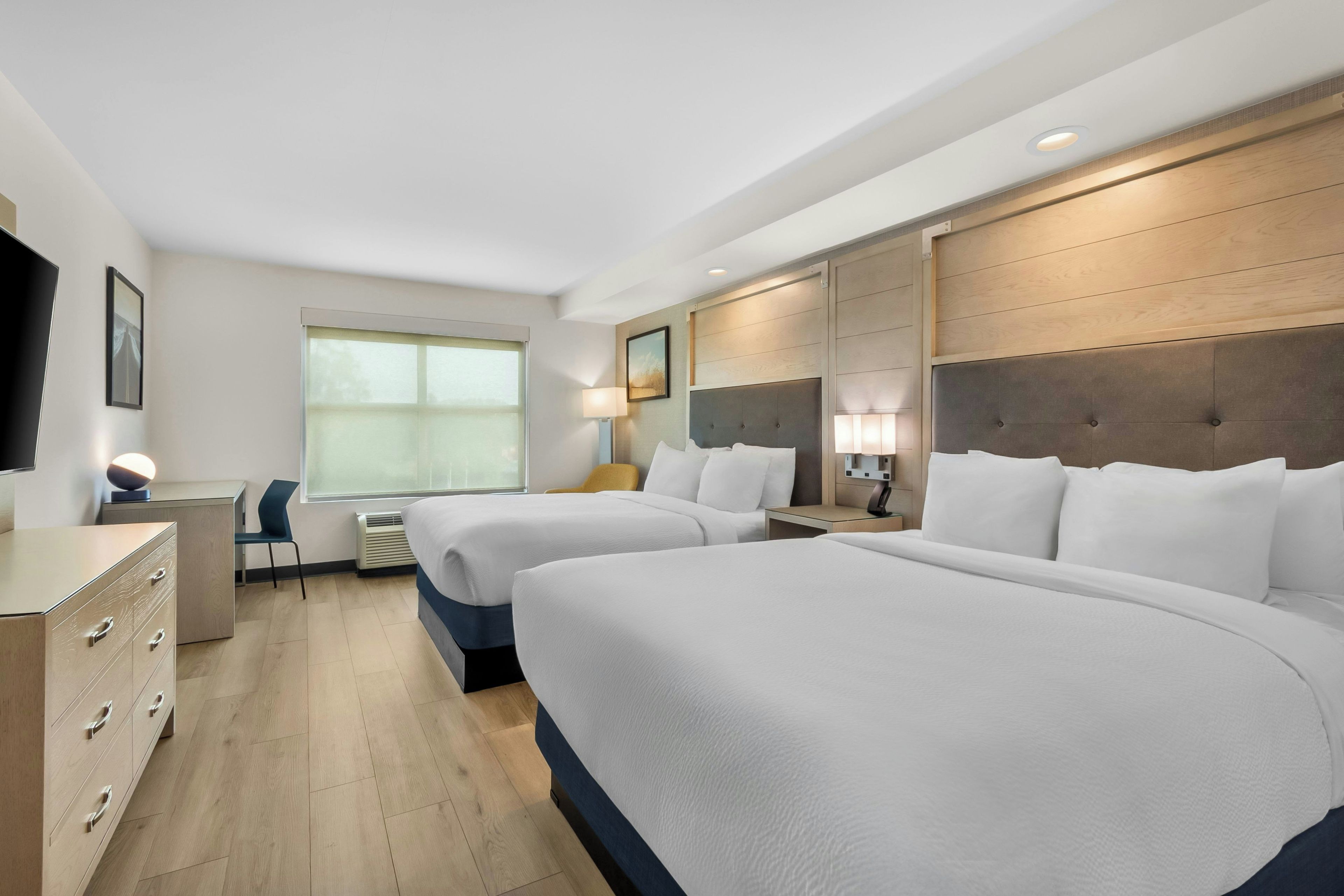 A hotel room with two beds, a desk, a chair, a TV, and a window.