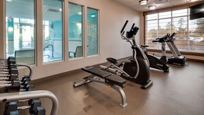 A gym with exercise equipment, large windows, and a view of parked cars outside.