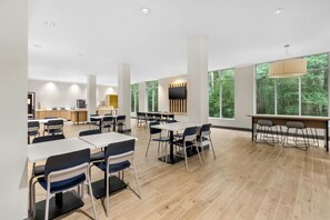 A modern dining area with tables and chairs, a kitchen in the background, and large windows with a forest view.