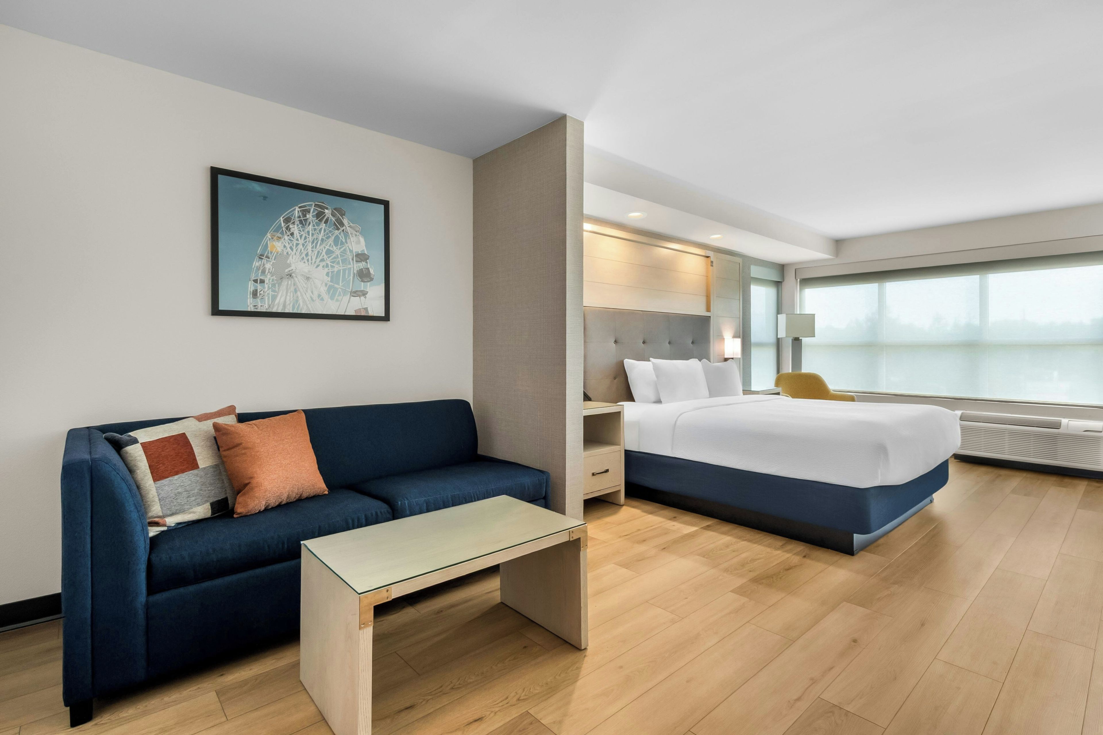 Una habitación de hotel con cama, sofá, una mesita, un buró, lámpara y un ventanal grande.