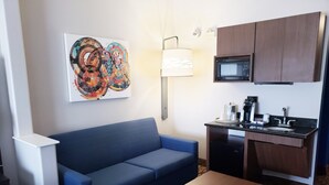 Et kompakt hotelværelse med en blå sofa, et lille træbord, et tekøkken med mikroovn og et billede på væggen.
