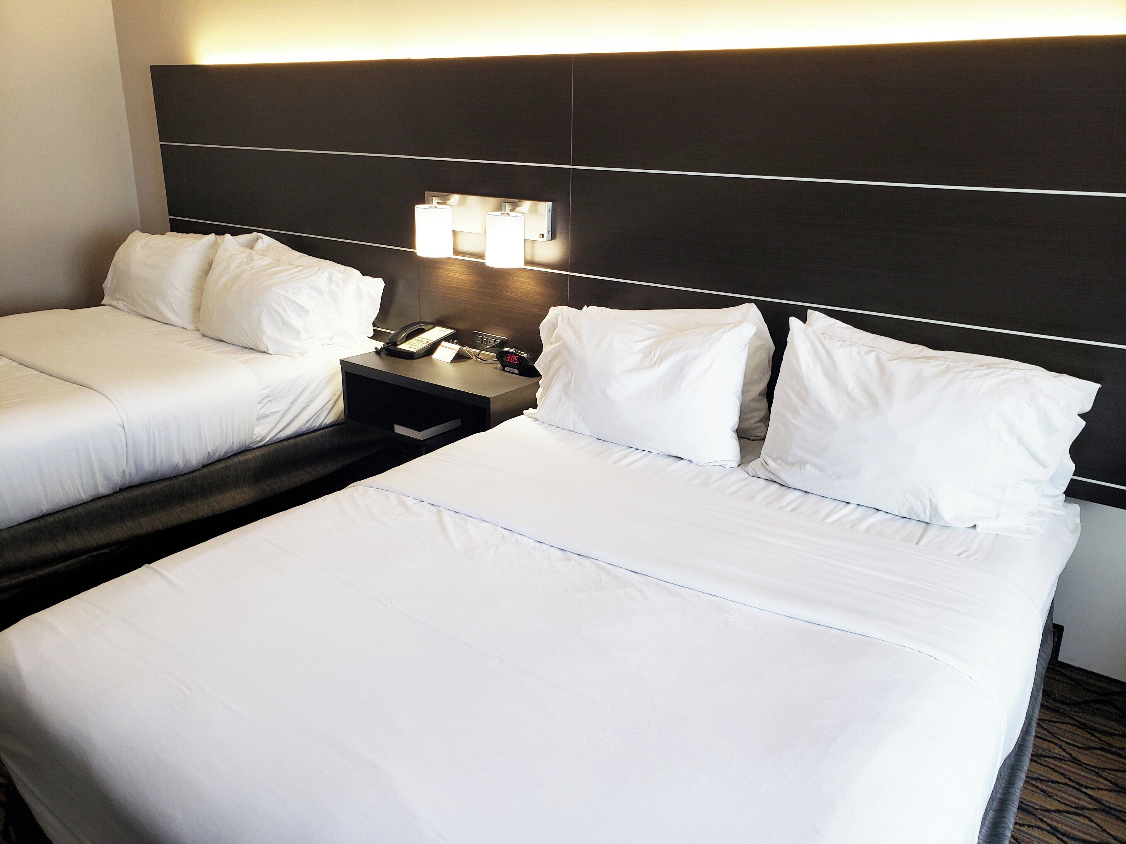 A hotel room with two beds, a nightstand with a phone and a clock, and a wall-mounted light fixture.