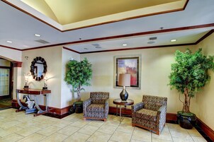 A lobby with two patterned armchairs, a small round table with a lamp, a potted plant, and a decorative mirror.