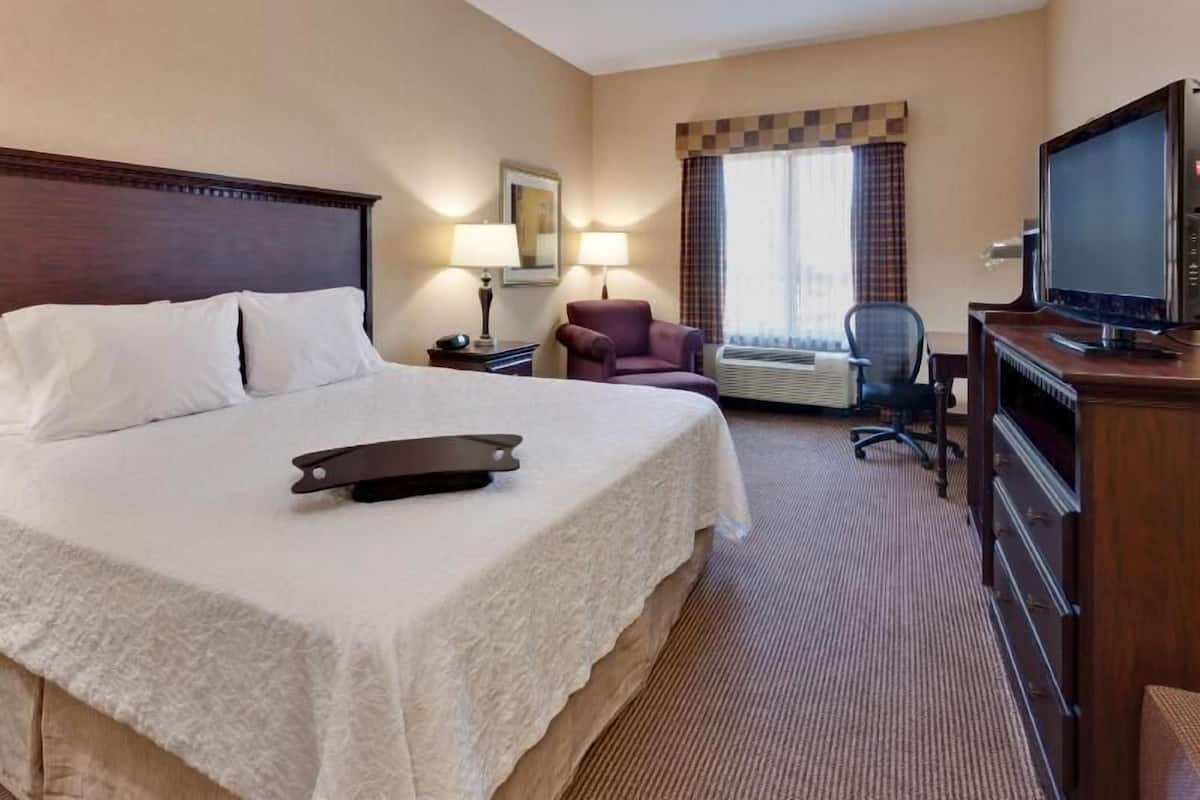 A hotel room with a large bed, a desk with a television, a chair, and an air conditioning unit.