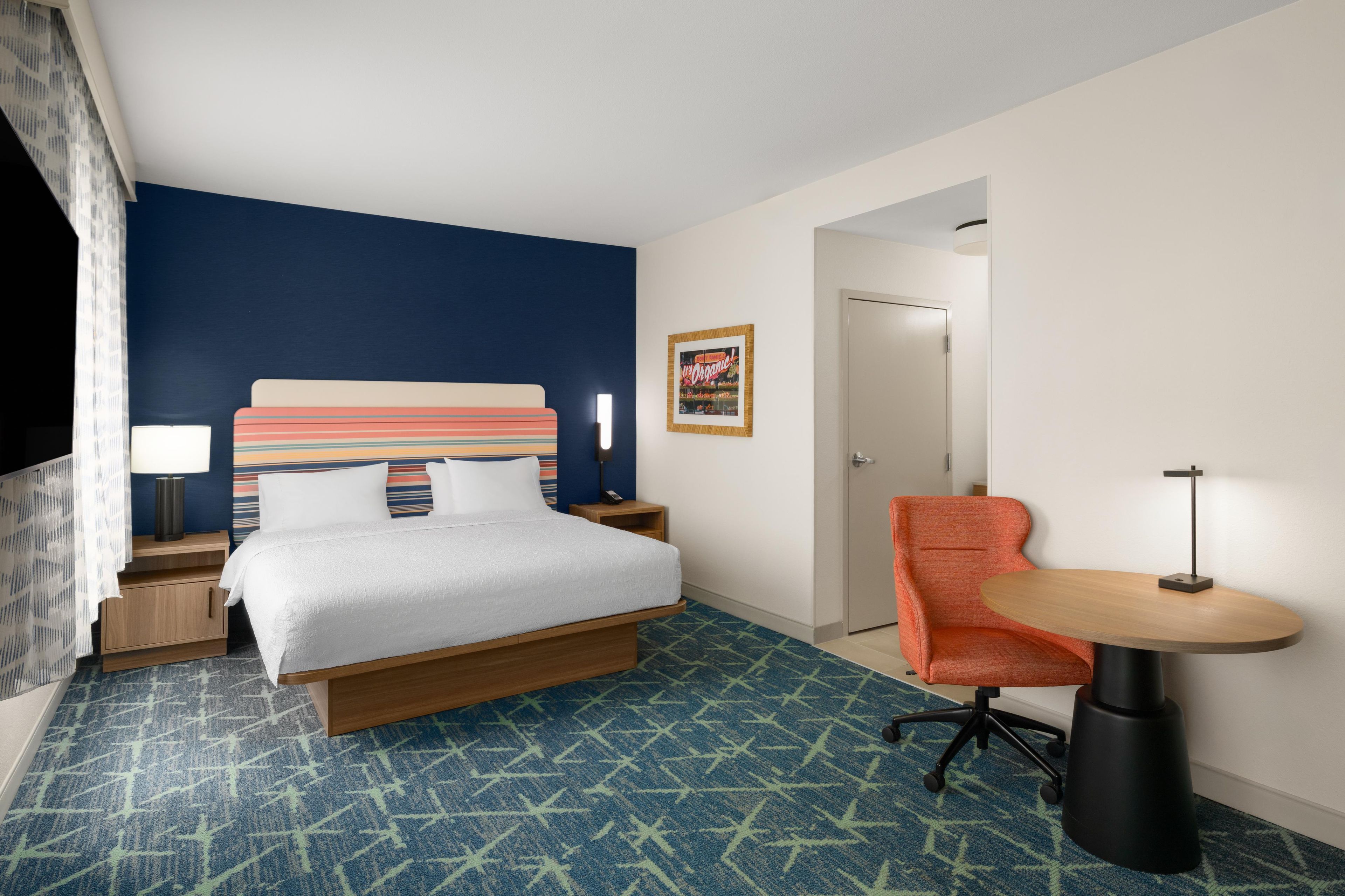 A hotel room with a bed, a desk, a chair, a lamp, and a door.