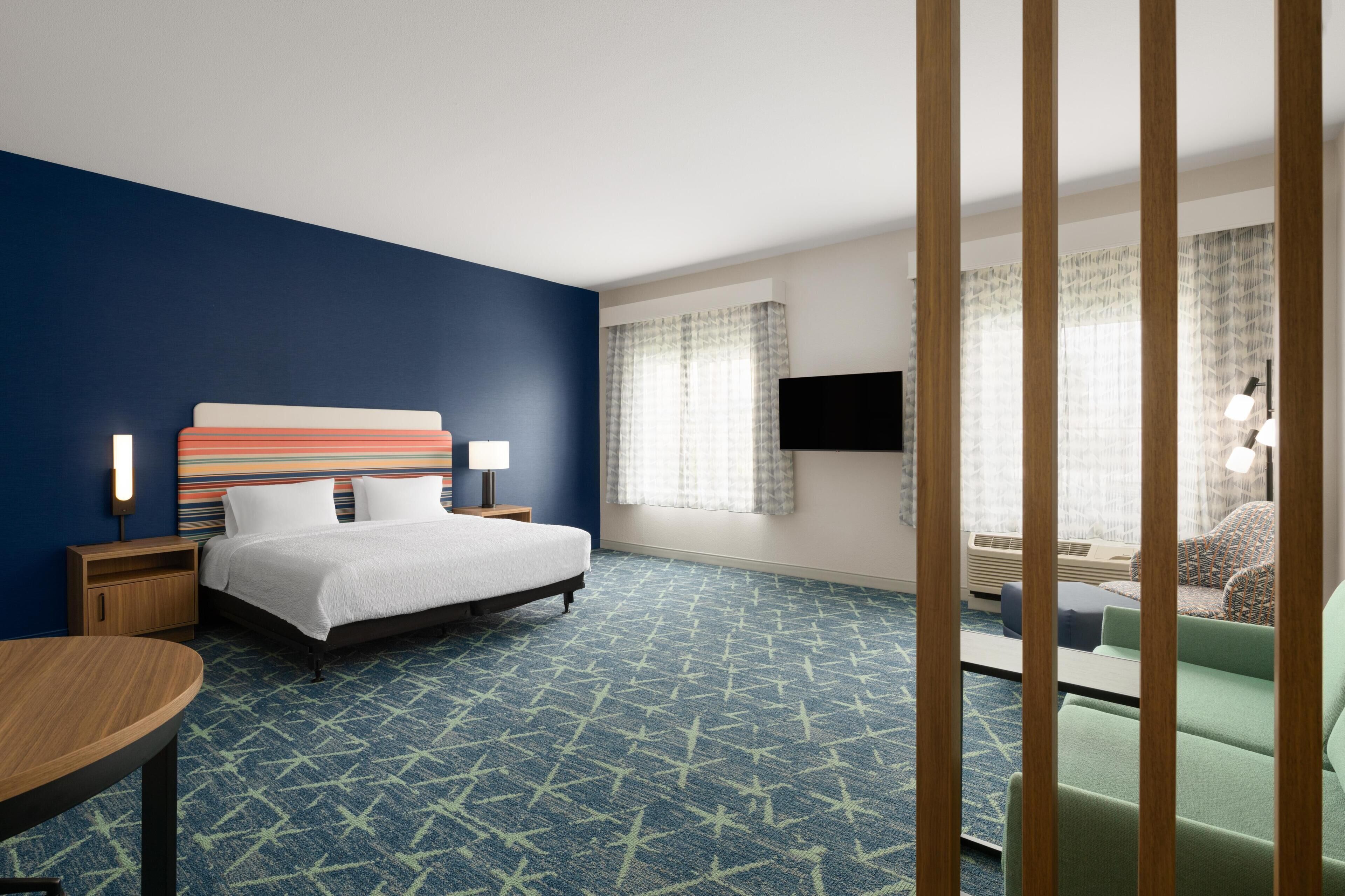 Quarto de hotel com cama, mesas de cabeceira, escrivaninha, cadeira, televisão e janela com cortinas.