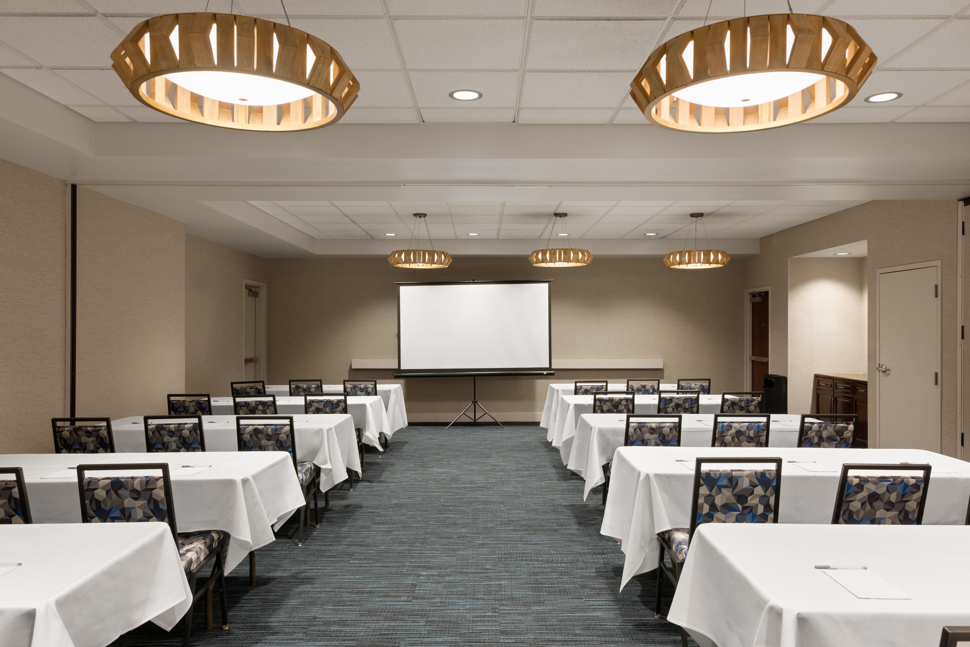 Un salón de conferencias con mesas, sillas, una pantalla de proyector e iluminación colgante.