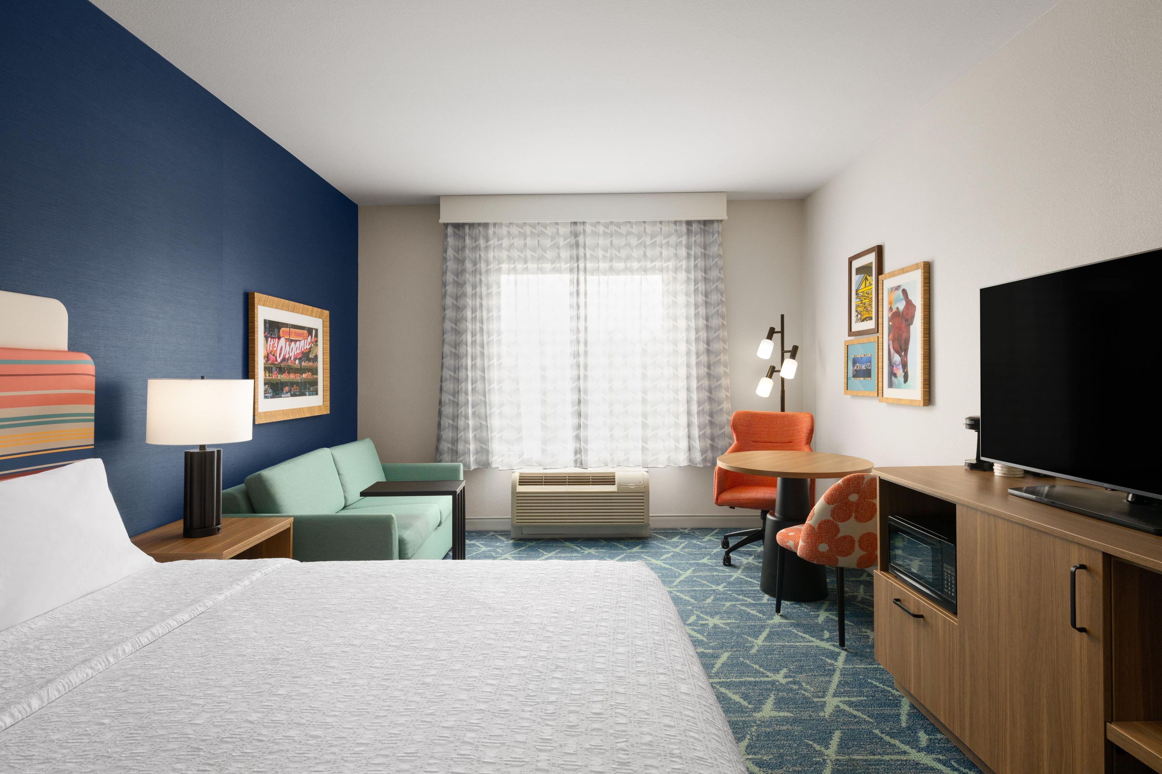 Quarto de hotel com cama, escrivaninha com televisão, cadeira, sofá, abajur e janela com cortinas.