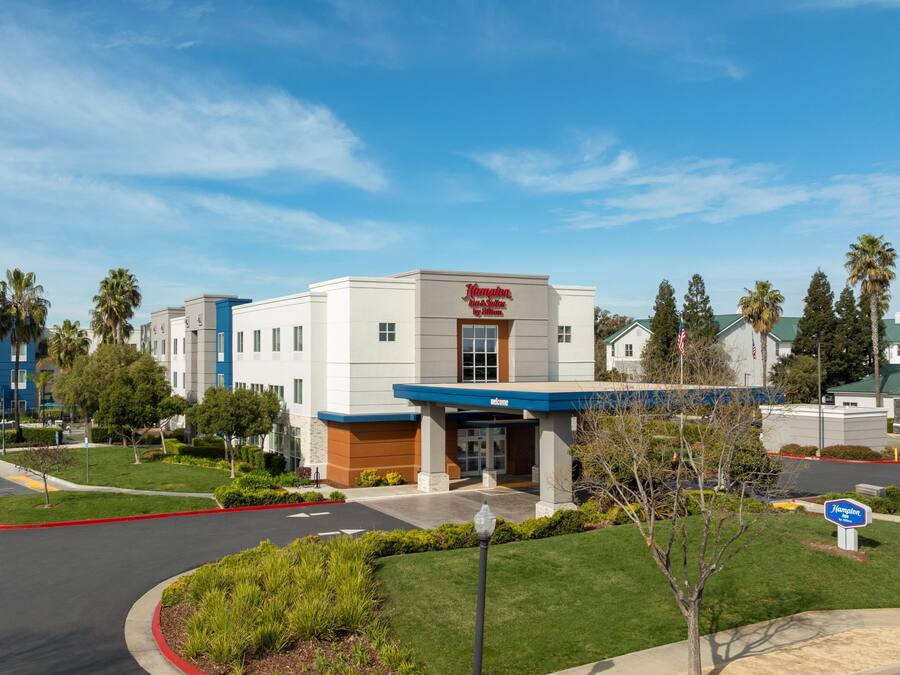 A Hampton Inn & Suites by Hilton hotel with a modern facade, a landscaped entrance, and a parking area.