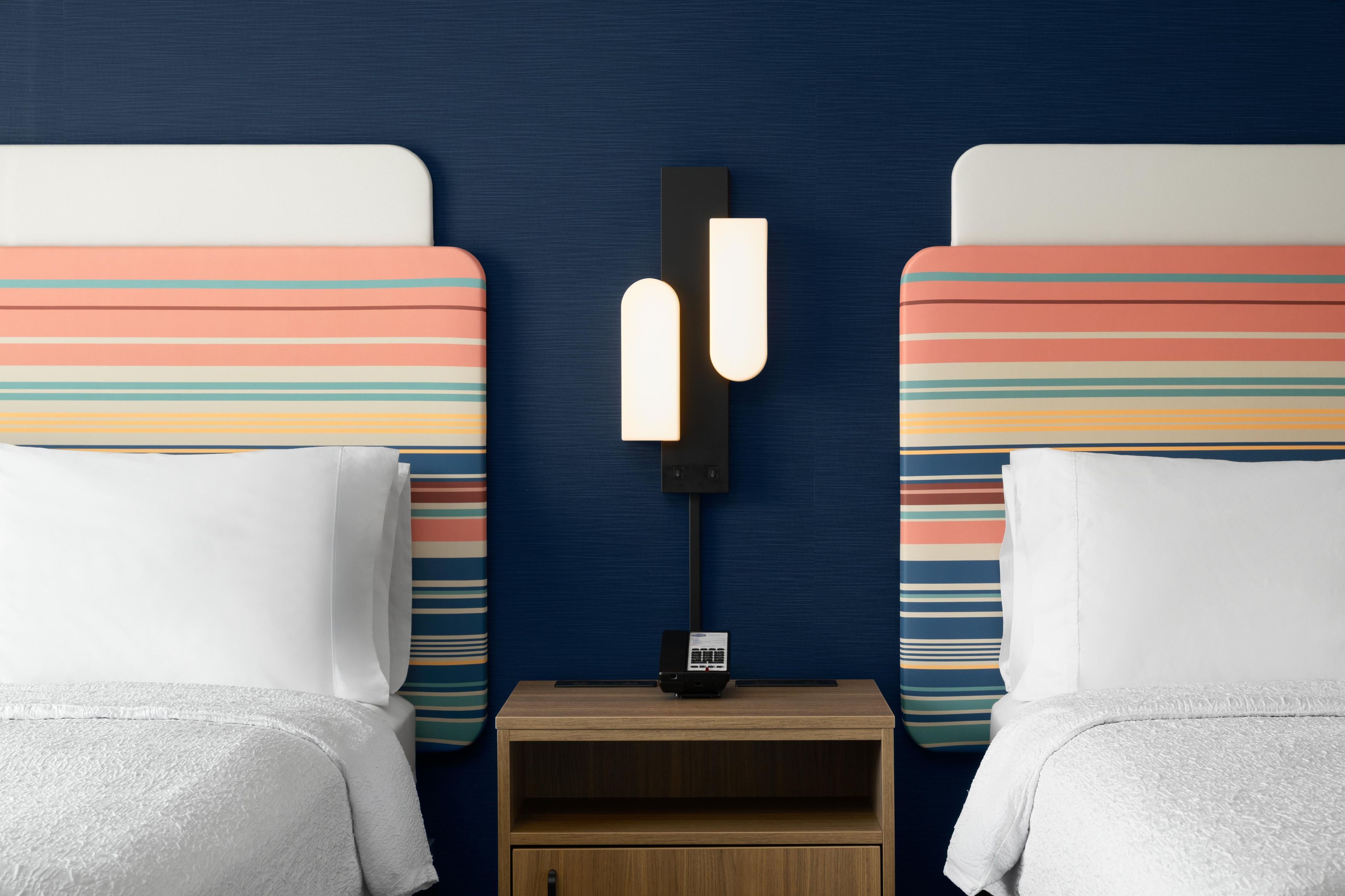 A hotel room with two beds, each with a striped headboard, a bedside table with a lamp, and a small drawer.