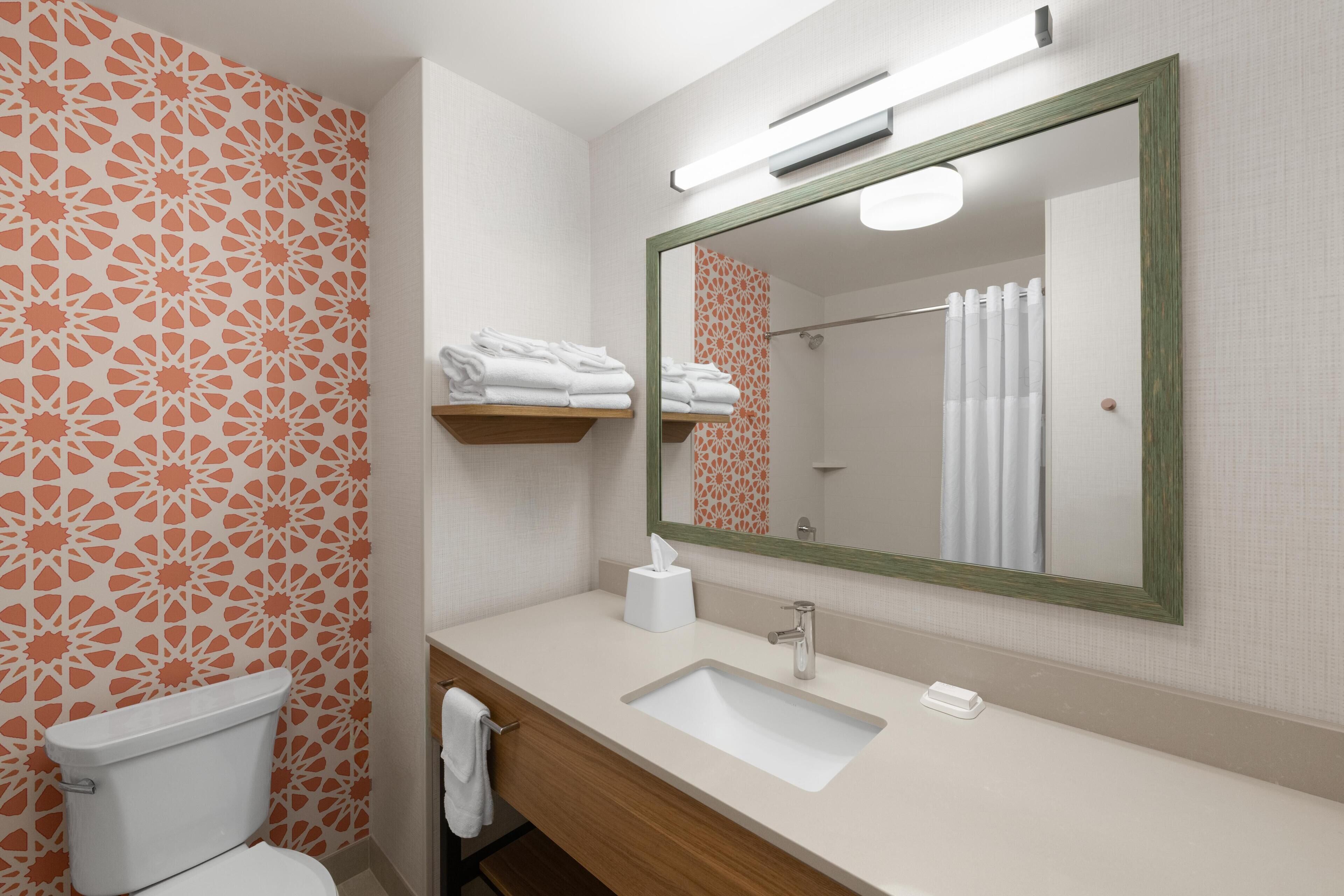 Un baño con inodoro, lavamanos y espejo. Hay toallas en un estante y una cortina de ducha.