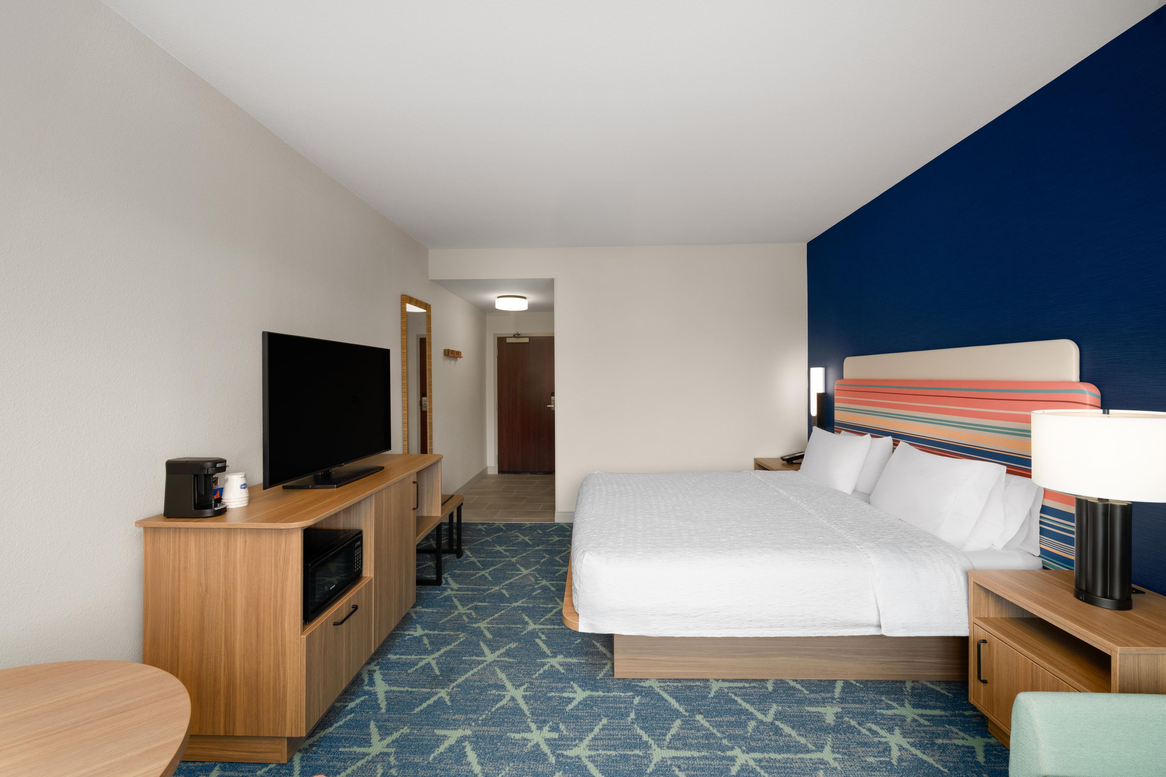 A hotel room with a bed, a TV, a desk with a coffee maker, and a bathroom door.