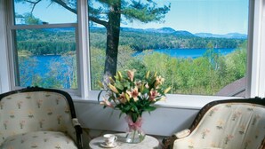 Una habitación con dos sillones tapizados, una mesita con un jarrón de flores y una ventana que ofrece vistas a un lago y un bosque.