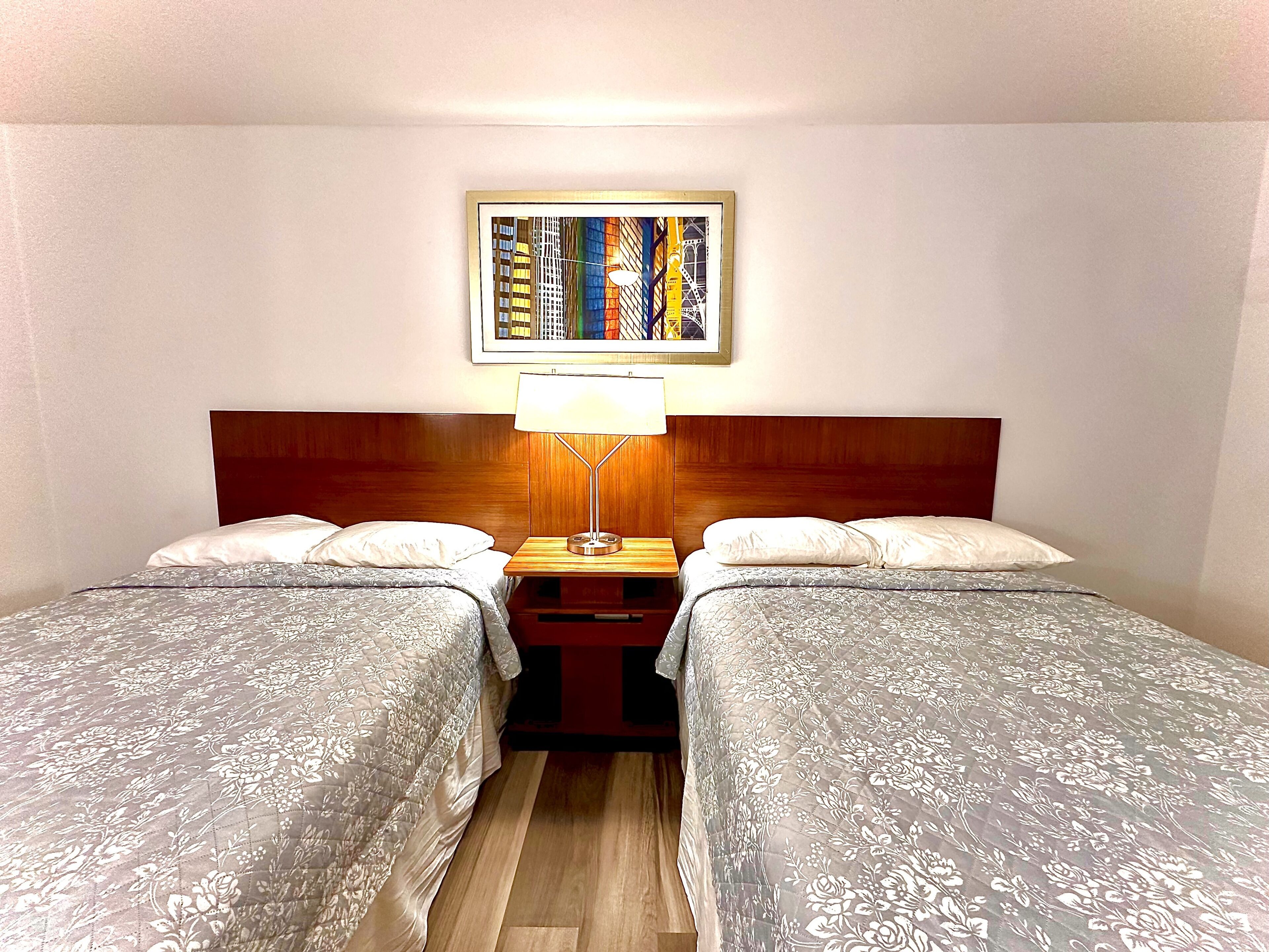 Habitación de hotel con dos camas, cabecera de madera, mesita de noche con lámpara y un cuadro enmarcado en la pared.