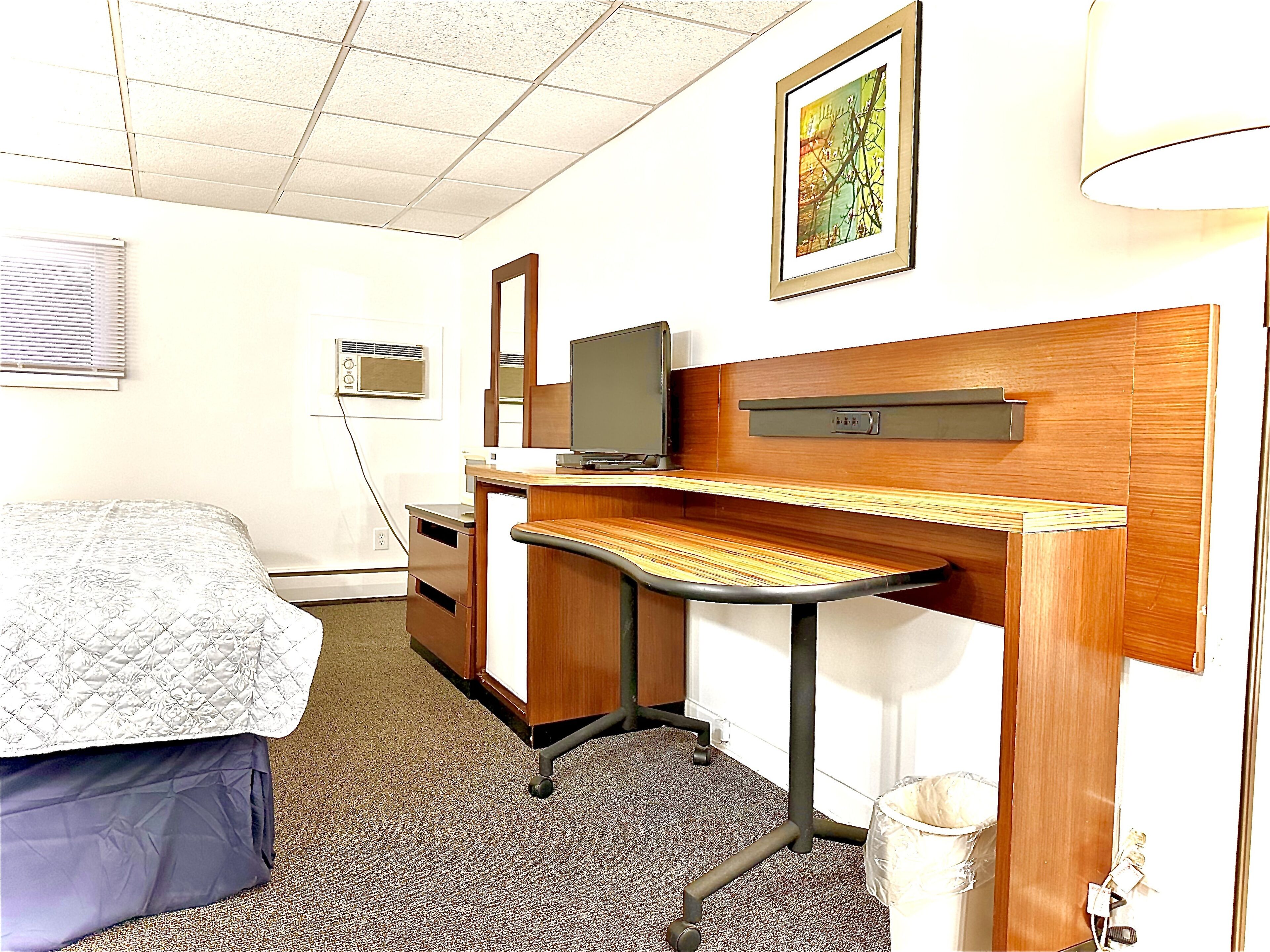 Une chambre d’hôtel équipée d’un lit, d’un bureau, d’une télévision, d’un miroir, d’une poubelle et d’un luminaire au plafond.