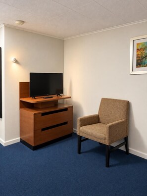 A room with a television on a wooden stand, a brown armchair, and a framed picture on the wall. - Crown Inn & Suites (Wellsville)